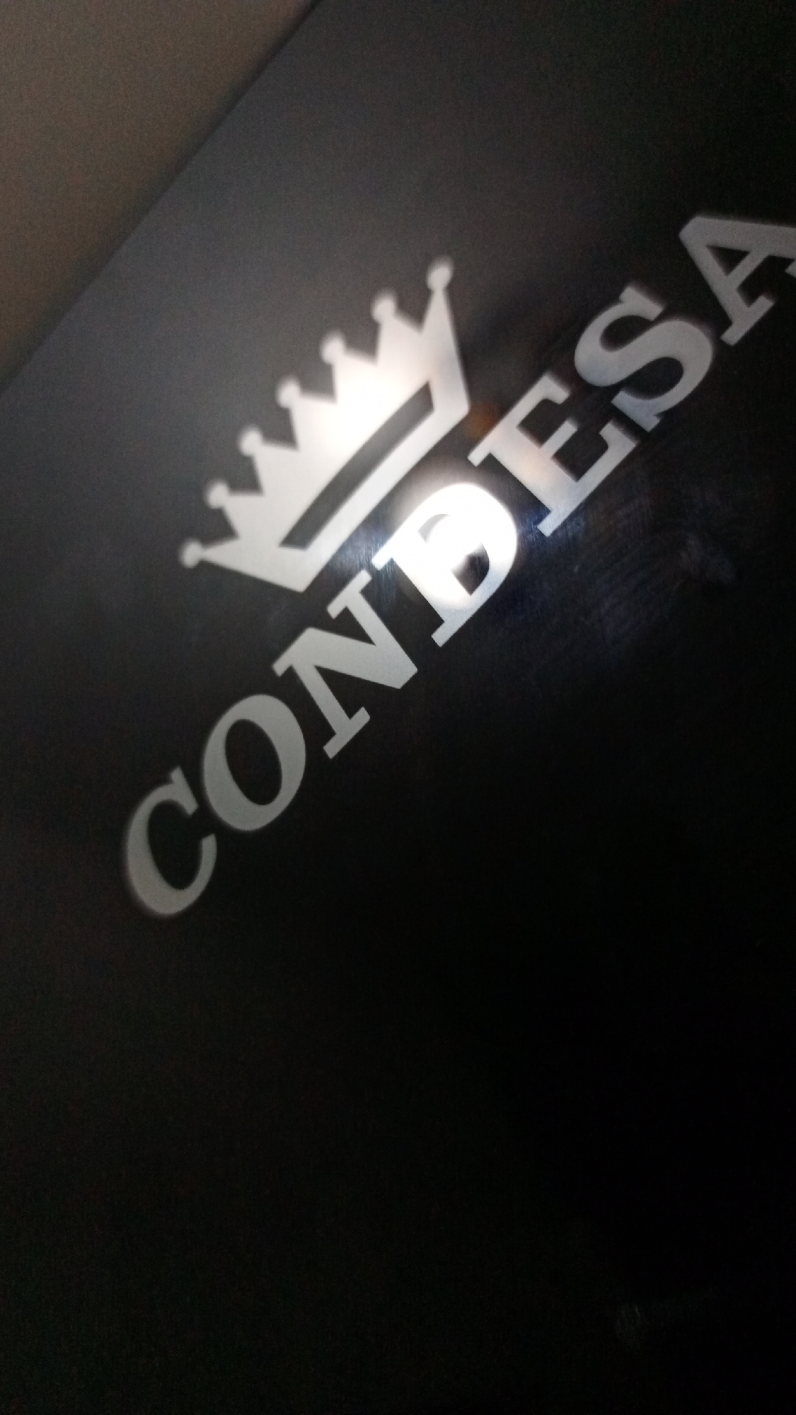 A dimly lit, slightly angled close-up of a black surface with the word "CONDESA" and a stylized crown logo printed in silver. The lighting is uneven, creating highlights and shadows. No people or specific location details are discernible in the image. The setting is indeterminable.