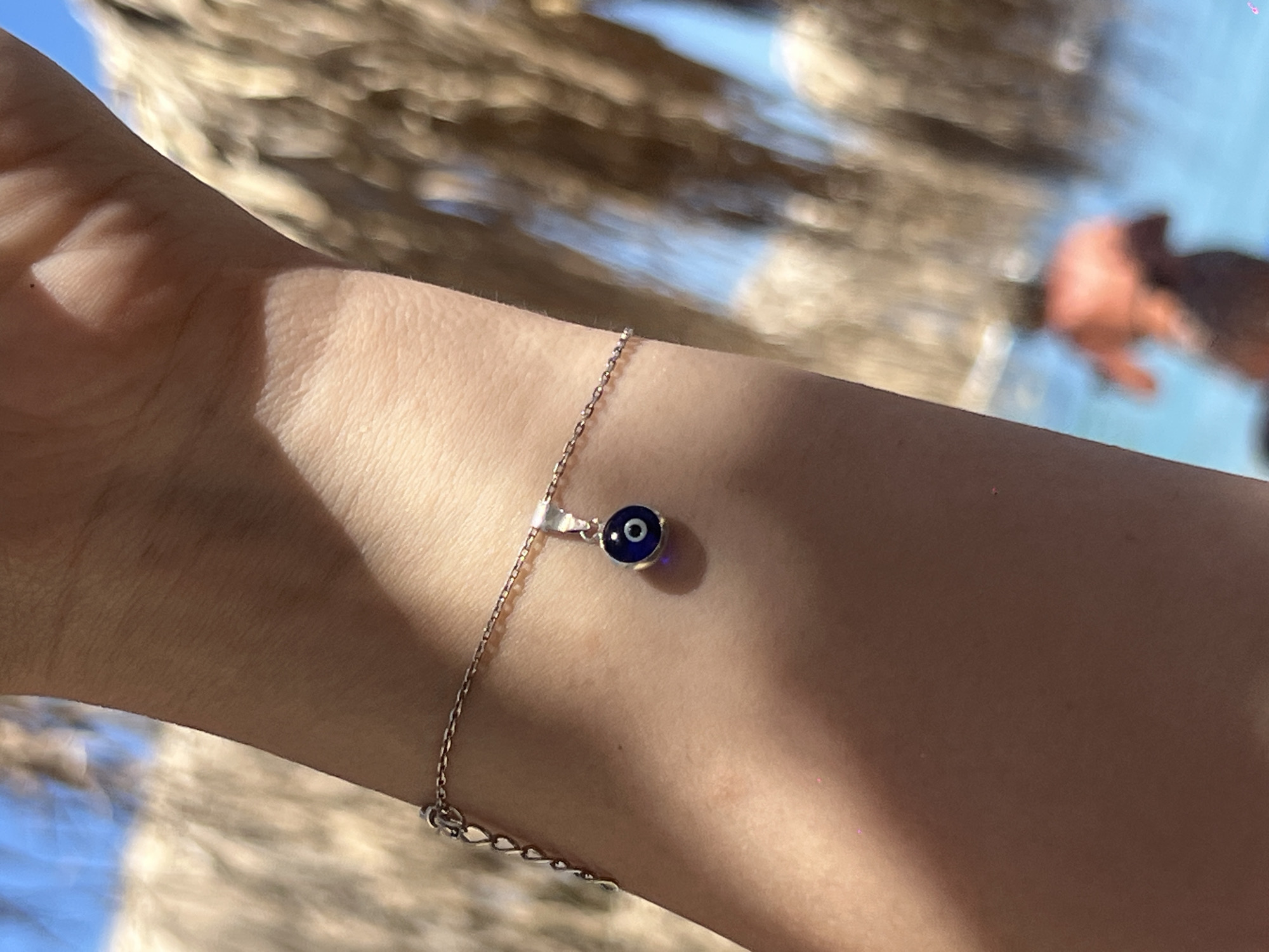 A close-up shot focuses on a person's wrist adorned with a delicate silver chain bracelet. The bracelet features a small, blue and white evil eye charm. The background is softly blurred, showing a bright blue sky with some white clouds and the dappled sunlight filtering through palm fronds, suggesting an outdoor setting. In the further background, a person in an orange garment is visible near a body of water. The location is identified as Achladias, Greece.
