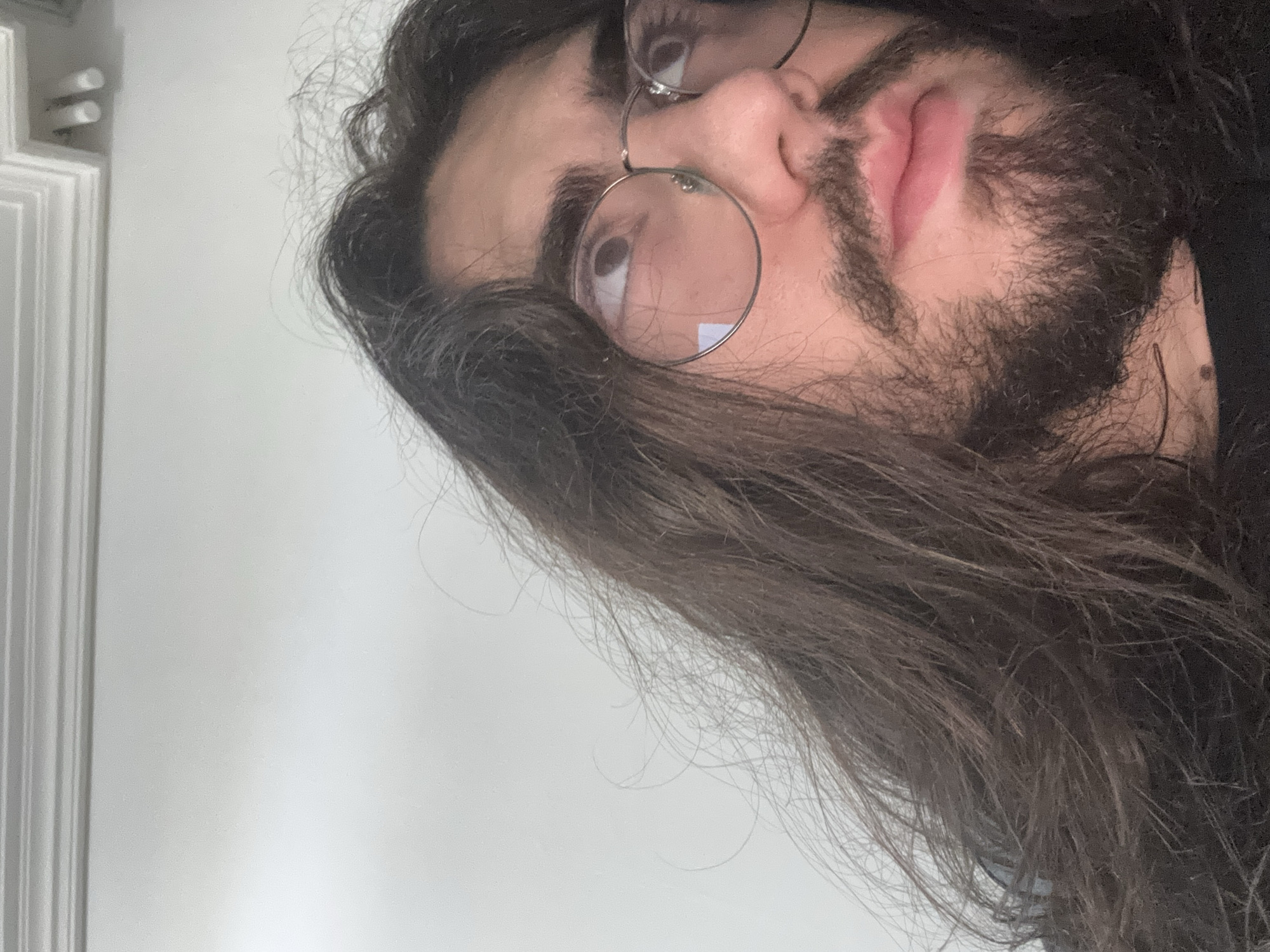 An extreme close-up, low-angle indoor shot captures a person with long, dark, wavy hair and a beard, wearing thin-rimmed glasses. The person's face is turned upward and to the left, with their gaze directed off-camera. The lighting appears to be from overhead, creating subtle reflections on the glasses. The background is a plain, light-colored wall, with a white decorative molding visible at the top of the frame.