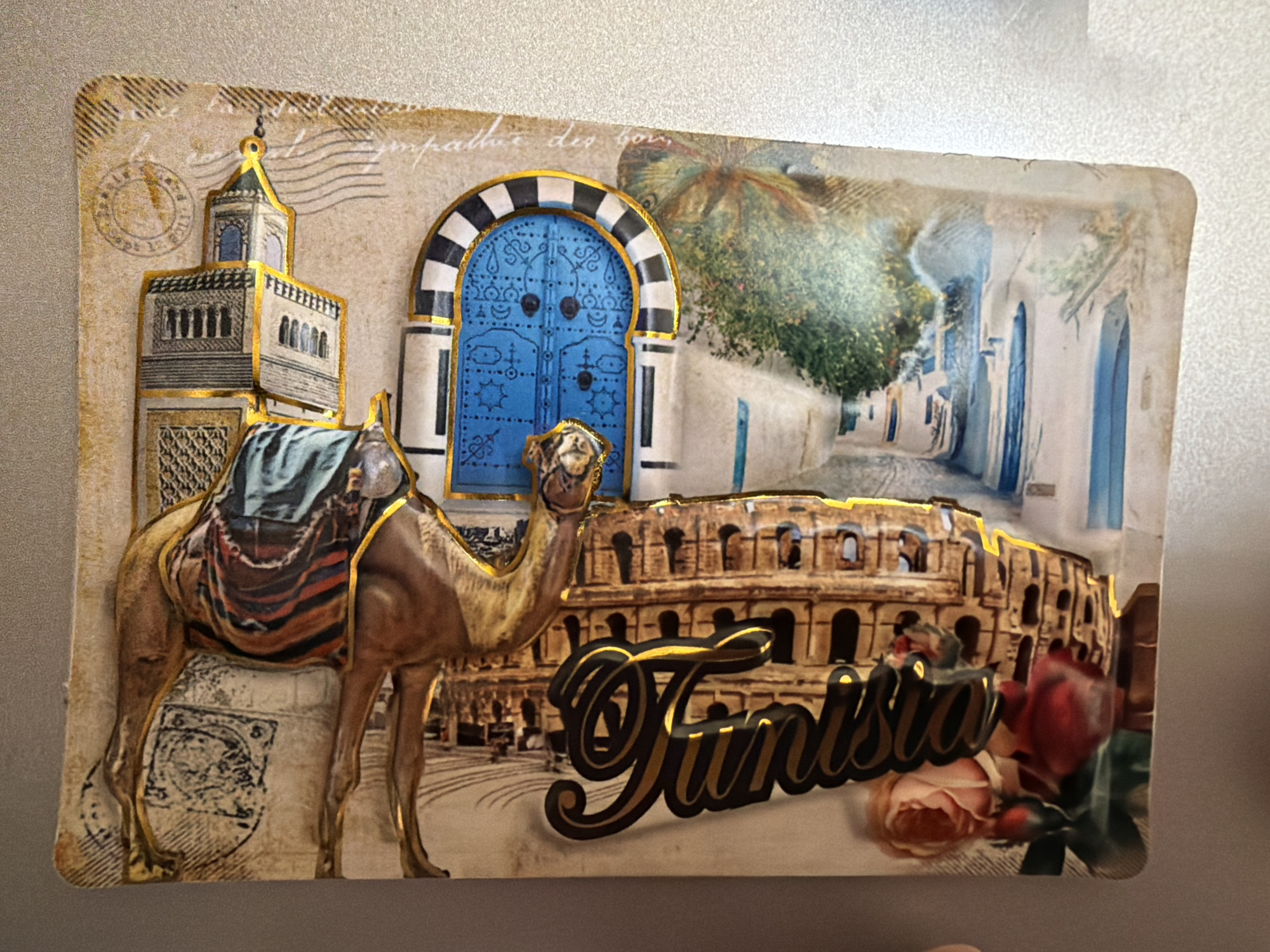This image is a close-up, slightly angled shot of a decorative souvenir or magnet, likely designed to represent Tunisia. The souvenir features a collage of iconic imagery associated with the country.
In the foreground, on the left, is a relief sculpture of a camel with a colorful saddlebag. Behind the camel, a stylized minaret of a mosque rises, adorned with gold trim. To the right of the minaret is a prominent, ornate blue arched doorway, also outlined in gold.
In the background, a depiction of the Colosseum in Rome is visible, suggesting a fusion of North African and Mediterranean historical themes. Overlapping the Colosseum is a street scene with white buildings and blue doors, characteristic of Mediterranean coastal towns. A palm tree is also visible in this background section.
The text "Tunisia" is prominently displayed in stylized, glossy black and gold lettering across the lower center of the souvenir. Red and pink roses are placed to the right of the text, adding a floral element. The overall background of the souvenir appears to be designed to resemble aged parchment or a vintage postcard, complete with faux postmarks and script.
The image does not depict the city of Pazardzhik, Bulgaria, or any elements associated with it.