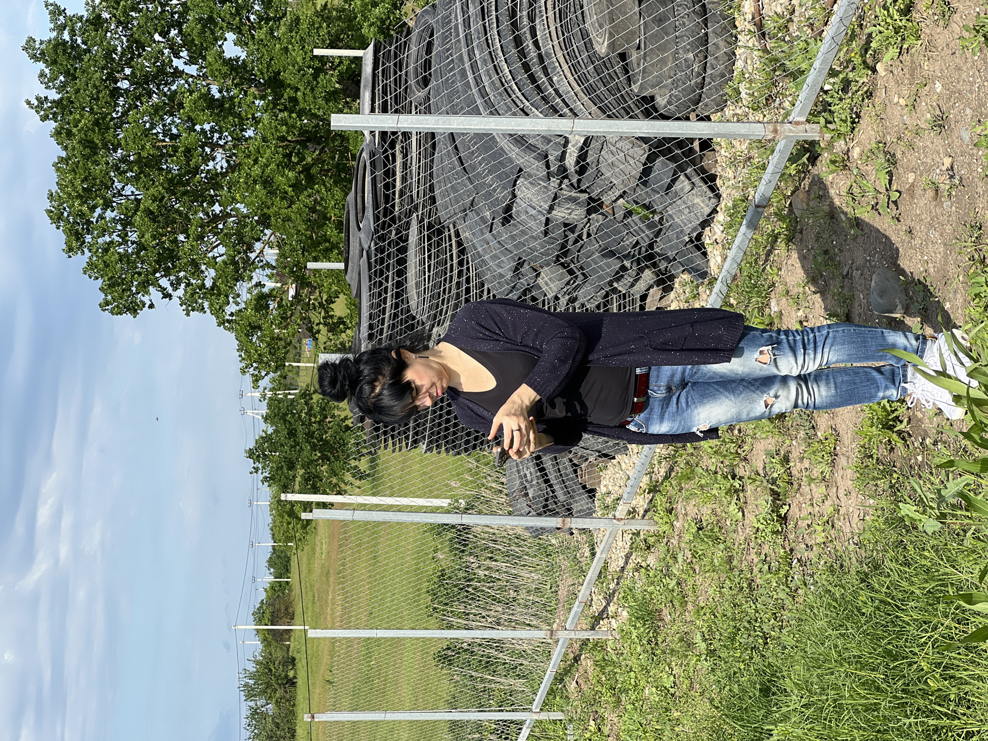A woman with dark hair styled in a bun stands in front of a chain-link fence in a grassy area. Behind the fence are several large black tires piled on top of each other. She is wearing a black shirt, a dark cardigan with sparkle details, ripped blue jeans, and white sneakers. Her head is tilted down, and she appears to be looking at something in her hands. The environment includes a field, trees, utility poles, and a blue sky with clouds in the background. The location appears to be outdoors in a rural or semi-rural area. While Tugurgoy, Russia is a potential location, this cannot be confirmed with certainty from the visual information provided.
