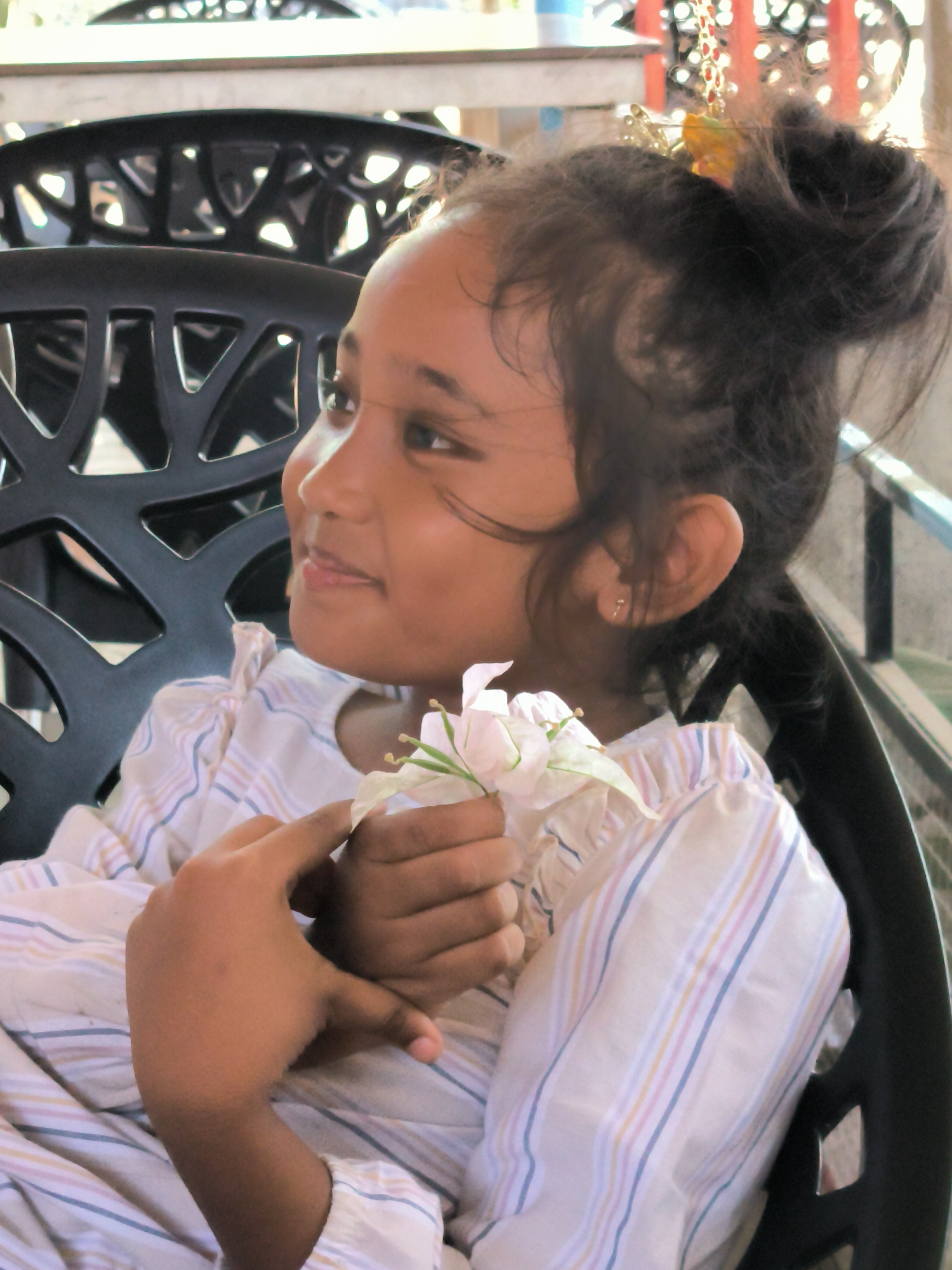 A young girl sits on a black plastic chair with an openwork design, holding a white flower. She is wearing a light-colored, striped shirt. Her hair is styled in a bun with a decorative hair accessory. The background includes other chairs and glimpses of a railing and greenery. The location is Matang, Indonesia.