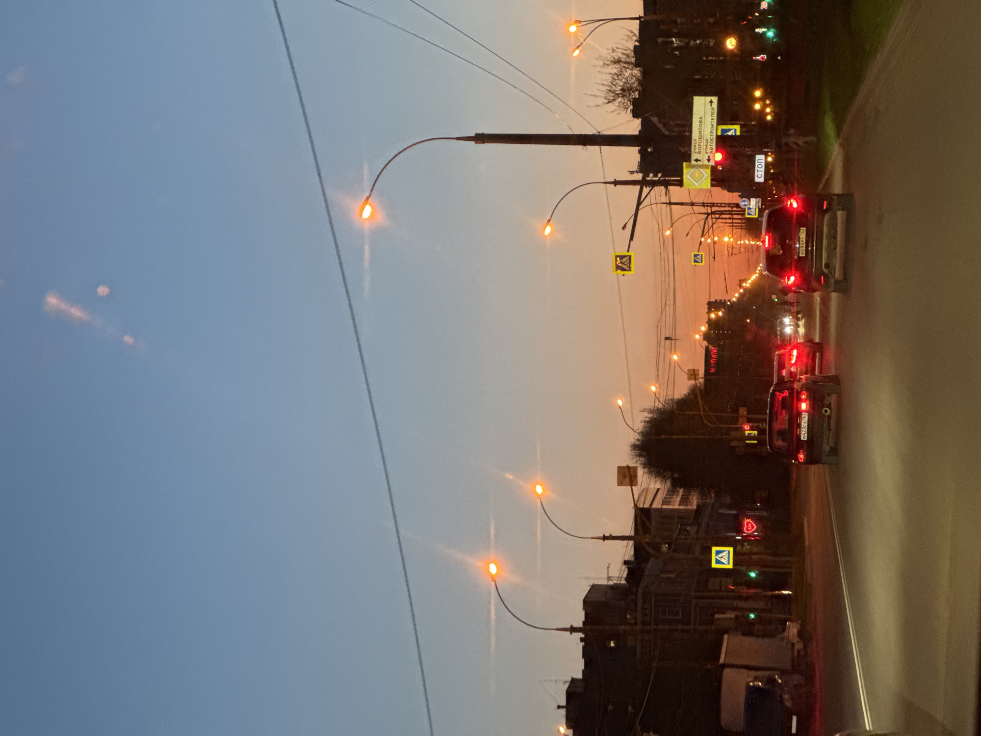 Here is a description of the image:
The image depicts a street scene at dusk in an urban area. The sky transitions from a light blue on the left to an orange glow near the horizon. Streetlights with curved arms illuminate the street, casting a warm light. The street itself appears to be a main thoroughfare with multiple lanes. Traffic is visible, including cars with their taillights illuminated. Street signs, including pedestrian crossing signs, are present along the roadside. Buildings line the street, silhouetted against the sky. Power lines stretch across the upper part of the frame. The road and sidewalk are visible with a pair of cars on the street. There are trees with green leaves to the right of the street. The location is in the city of Unknown, Unknown.