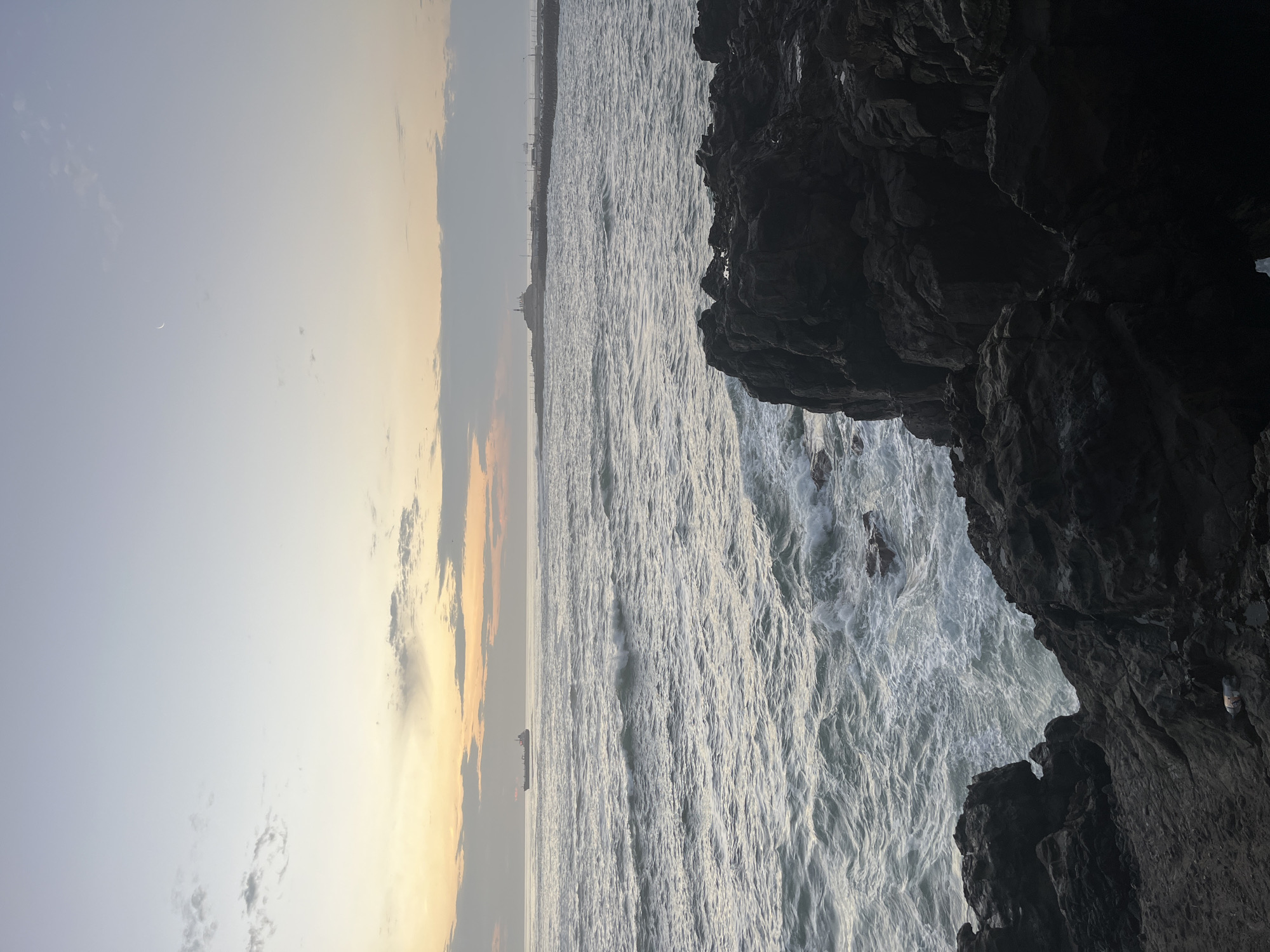 The image depicts a coastal scene in Arica, Chile at twilight. The sky ranges from pale blue to hues of orange and grey with some clouds. A crescent moon is faintly visible in the sky. Below, the ocean appears choppy and grey with whitecaps. In the distance, a cargo ship and a few distant coastal structures are visible. Large, dark, jagged rocks dominate the right side of the frame. White waves crash against these rocks.