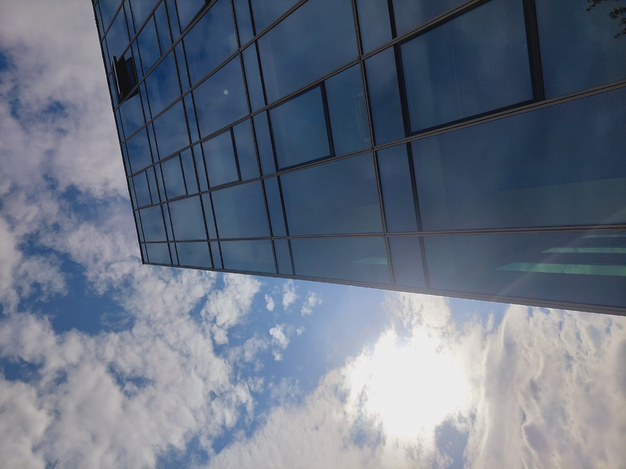 An abstract, low-angle shot captures a modern glass-paneled building against a backdrop of a partly cloudy blue sky. The sun is bright, creating lens flares and reflections on the building's dark, tinted windows. One window near the top of the building is slightly ajar. The geometric lines of the building's facade dominate the right side of the frame, while the expansive sky with scattered white clouds fills the left. There are no people or visible signs of activity within or around the building. The location is a city, though no specific landmarks or identifying features of Sofia, Bulgaria, are discernible in the image itself.