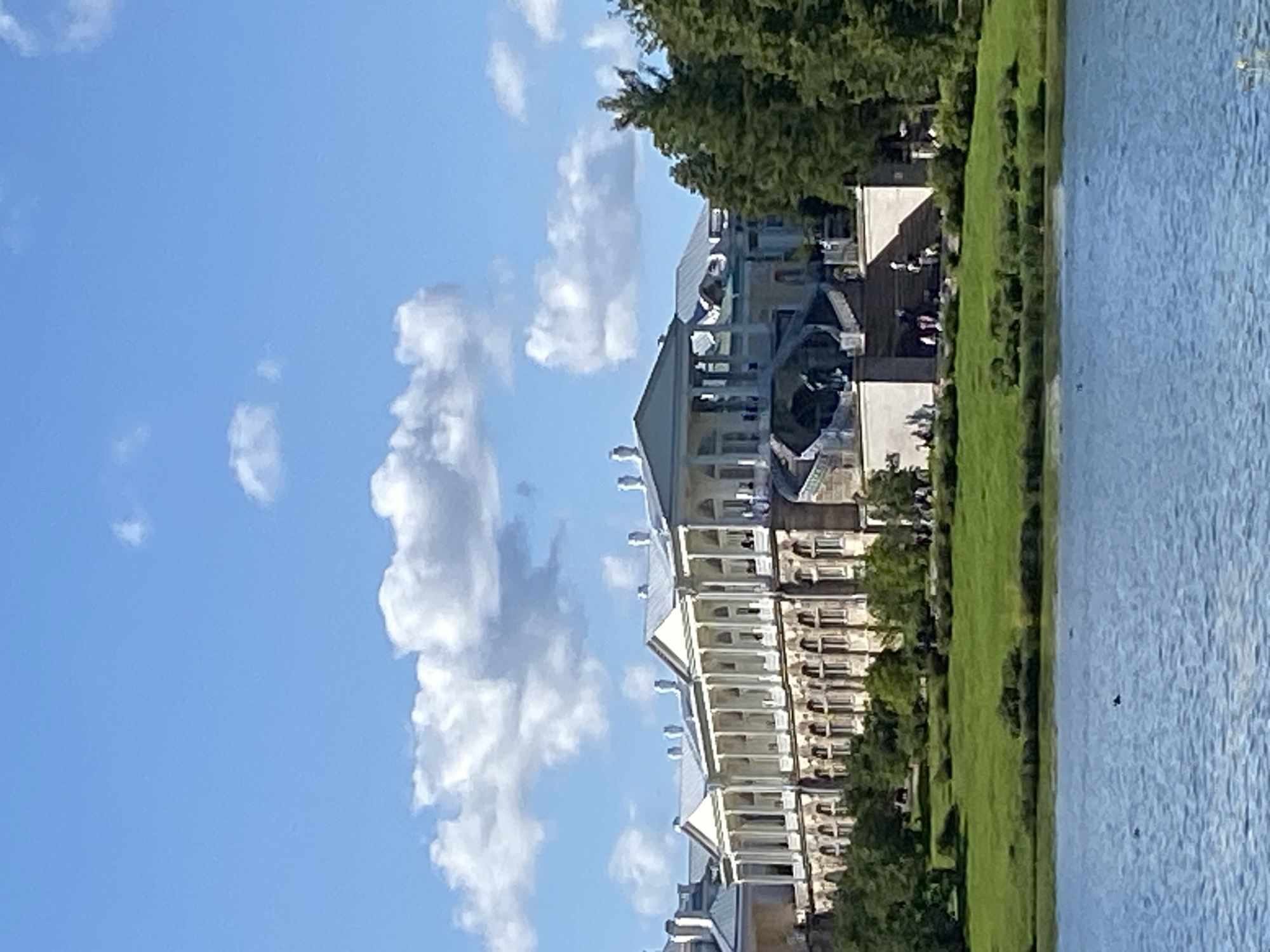 The image depicts a wide, panoramic view of the Catherine Palace in Pushkin, Russia, on a bright, sunny day. The palace, a grand Baroque structure, dominates the middle ground, characterized by its long, white facade, numerous columns, arched windows, and mansard roofs. A prominent, wide staircase leads up to the central part of the palace, and several people are visible ascending and standing on it. The palace is surrounded by lush green lawns and mature trees, with a body of calm, blue water in the foreground. The sky is a clear blue, dotted with fluffy white clouds. The overall scene conveys a sense of historical grandeur and natural beauty.