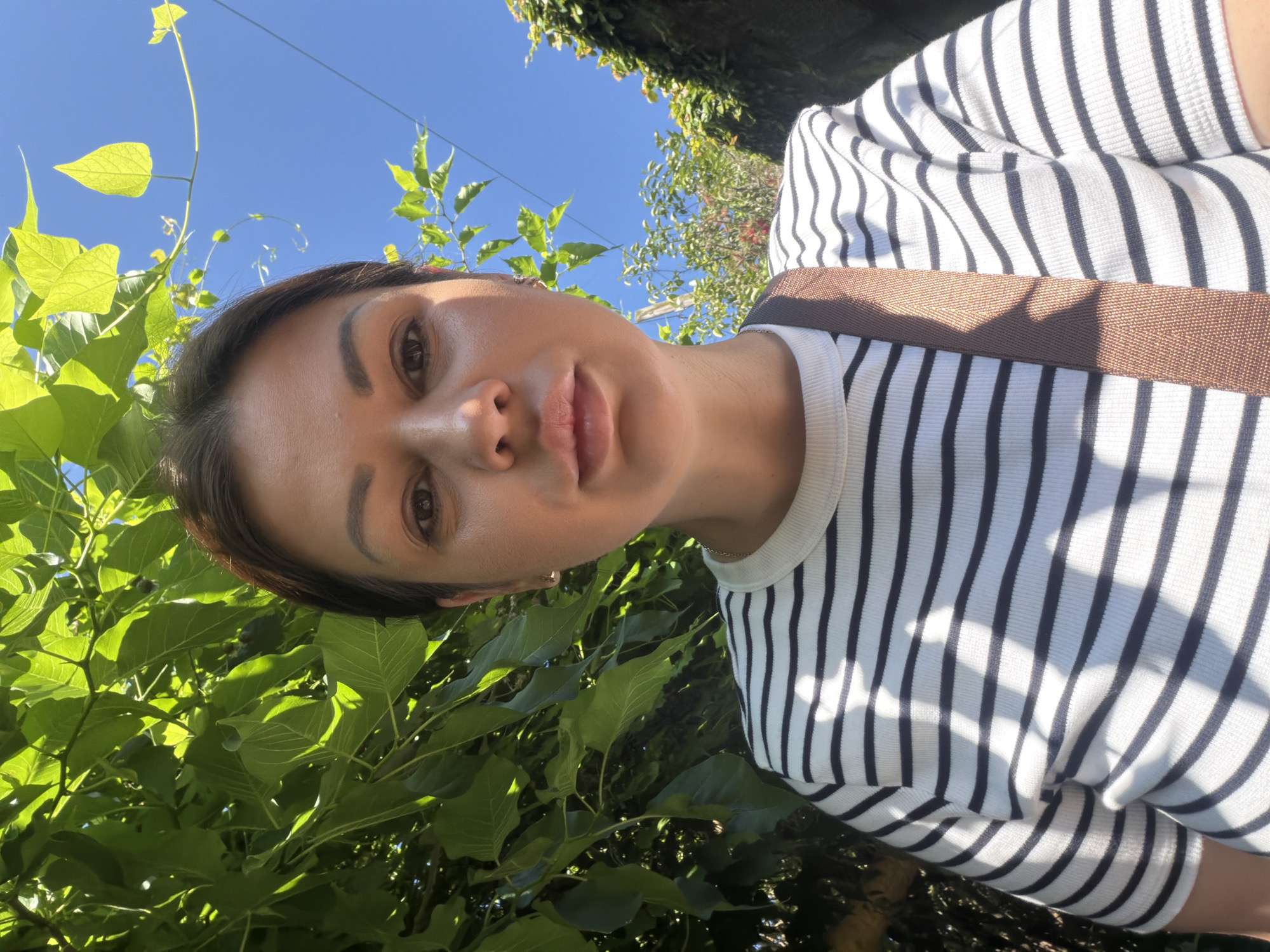 A medium-shot, waist-up, selfie-style photograph shows a woman standing outdoors in what appears to be a park or garden, possibly in Sochi, Russia, given the lush greenery and bright blue sky. The woman is facing the camera, and her head and upper torso are visible. She has dark hair pulled back, and her facial expression is neutral. She is wearing a white t-shirt with black horizontal stripes and a brown strap from a bag is visible across her chest. The background is dominated by vibrant green foliage, with dappled sunlight filtering through the leaves. The bright blue sky is visible above the greenery, suggesting it is a sunny day. The overall scene conveys a casual, outdoor setting.