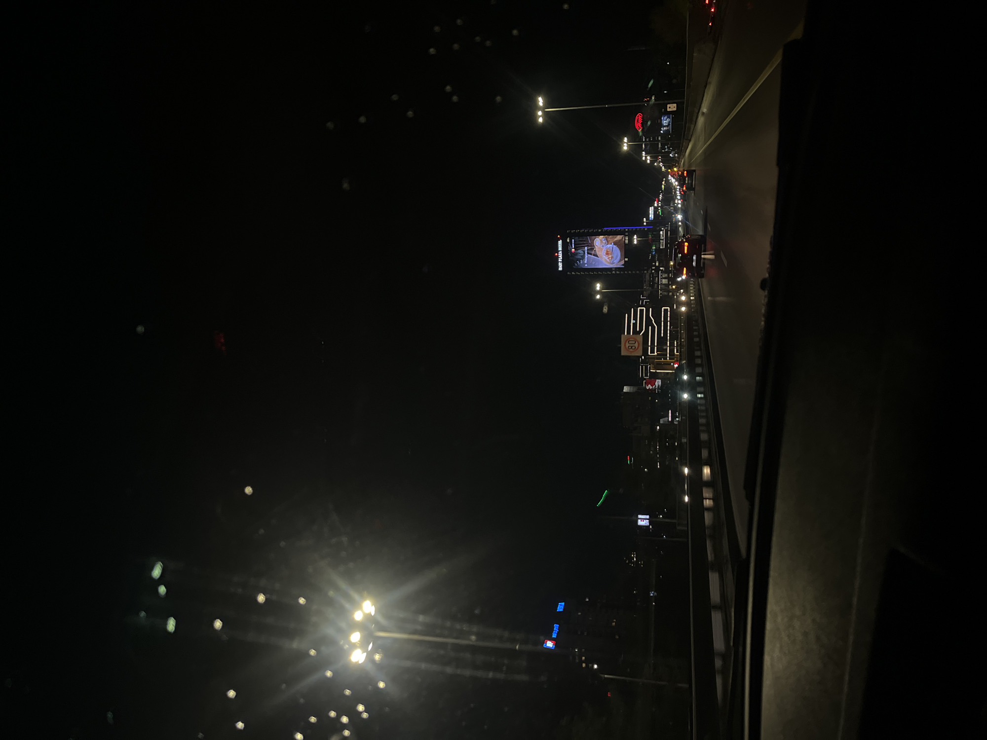 This is a nighttime, elevated view of a busy urban highway in Sofia, Bulgaria. The image is dominated by the darkness of the night sky and the vibrant artificial illumination of the road and roadside structures.
A multi-lane highway runs diagonally through the right side of the frame, filled with vehicles indicated by their illuminated headlights and taillights. The road surface reflects the bright lights, suggesting a wet or reflective surface.
Along the right side of the highway, a series of buildings and structures are brightly lit by commercial signs and streetlights. A prominent, tall building features a large digital billboard displaying an image of food and the text "EAST PLAZA HOTEL". Below it, a speed limit sign clearly shows "80". Further down the road, other illuminated signs are visible, including a blue sign with the word "VISA" and potentially "VIVA", and a distant red Coca-Cola logo. Numerous streetlights cast bright, starburst-like glares across the scene, contributing to the busy, energetic atmosphere.
The left side of the image is largely taken up by the dark night sky, with scattered, out-of-focus lights appearing as bokeh, possibly reflections or distant city lights. The immediate foreground on the far right shows a dark, solid structure, indicating the photo was likely taken from inside a vehicle or from an adjacent overpass or building. The scene captures the constant movement and commercial activity of a city highway after dark.