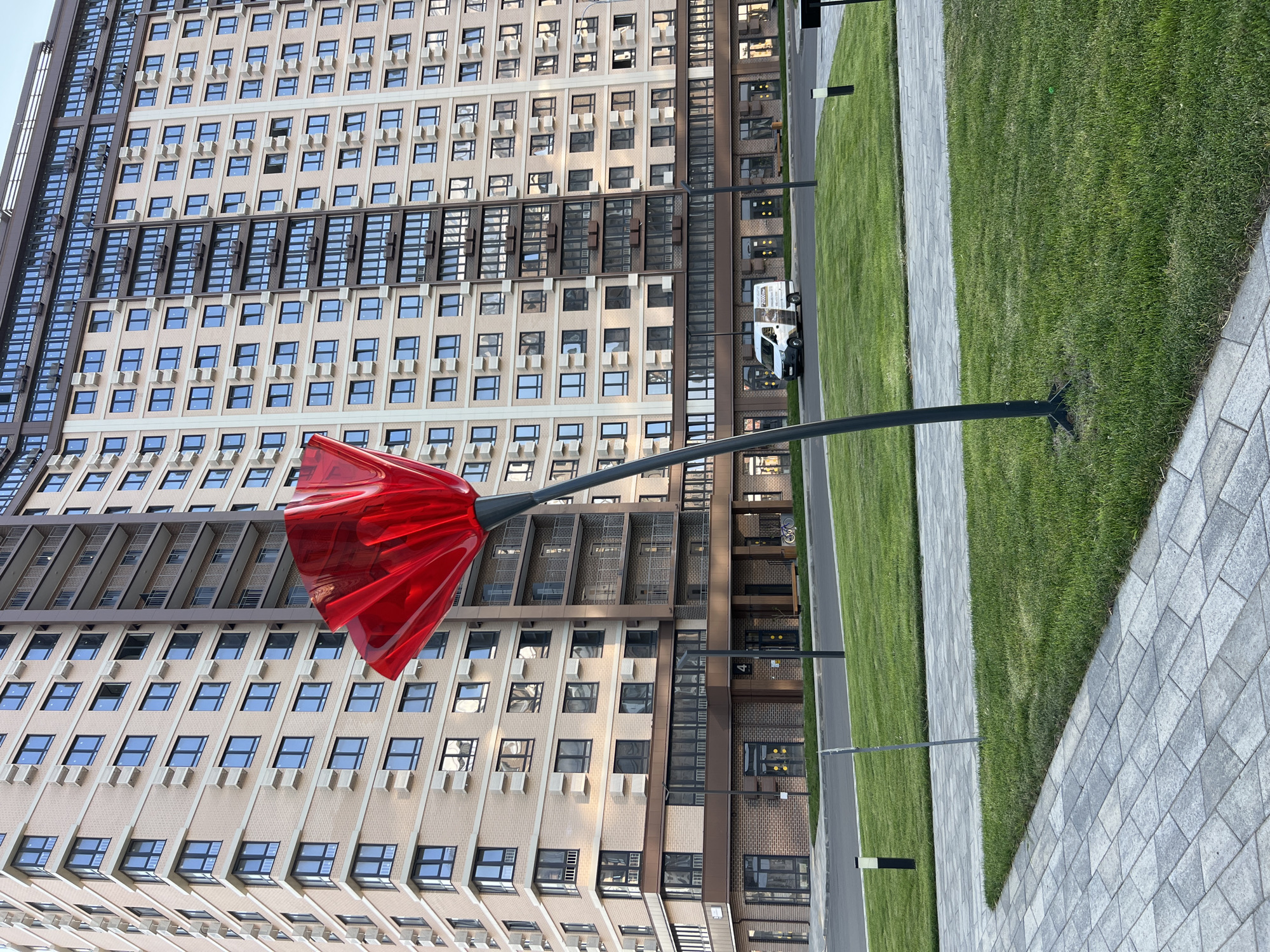 A large, modern residential building with a beige facade and numerous windows is visible. In the foreground, a tall, slender sculpture resembling a red poppy with a curved stem stands on a patch of green grass. A paved walkway runs alongside the grass. A white delivery van is parked on the street in front of the building's ground floor, which features storefronts. The scene suggests a new urban development.