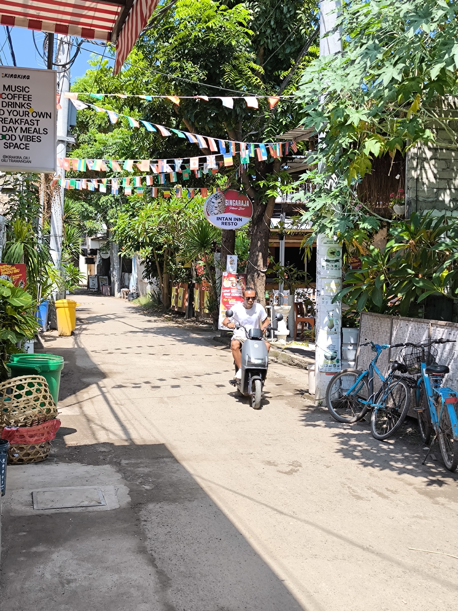 The image captures a street in Gili Trawangan, Indonesia, during the daytime. A man is riding a grey scooter along the street. The street is lined with buildings, trees, and plants. Bicycles are parked against a wall. Overhead are strings of colorful flags and banners, one for "KIRAKIRA" and "SINGARAJA." Trashcans and baskets are on the left side of the street. The street surface is asphalt. Shadows from the trees and buildings are cast on the street.