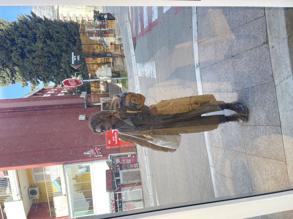 An individual is captured in a full-body reflection selfie taken in a large storefront window on a sunny day in Budva, Montenegro. The person stands centered, facing forward, holding a smartphone with their right hand to take the photograph. They possess medium-length wavy brown hair and wear dark sunglasses, a long olive-green trench coat with a belted waist, dark trousers, and dark combat boots. A white canvas tote bag is slung over their left shoulder. A small, orange, stylized mask-like charm hangs from the right hip area.
The reflection reveals a street scene across from the window. Directly opposite is a multi-story building, primarily dark red/maroon, featuring balconies and an air conditioning unit. Adjacent to it, another section of a building appears yellow. Visible signage includes "MTA BANKOMAT" in red lettering, confirming a financial service, and a prominent red octagonal "STOP" sign. A street sign with a left-pointing arrow indicates "DOWNTOWN 200m" (reversed in reflection). A tall, dense evergreen tree stands to the right, behind which distant mountains are visible under a clear blue sky. The street features a pedestrian crosswalk with white and red markings, and a motorcycle is parked near a storefront. The ground beneath the subject is a grey tiled sidewalk.