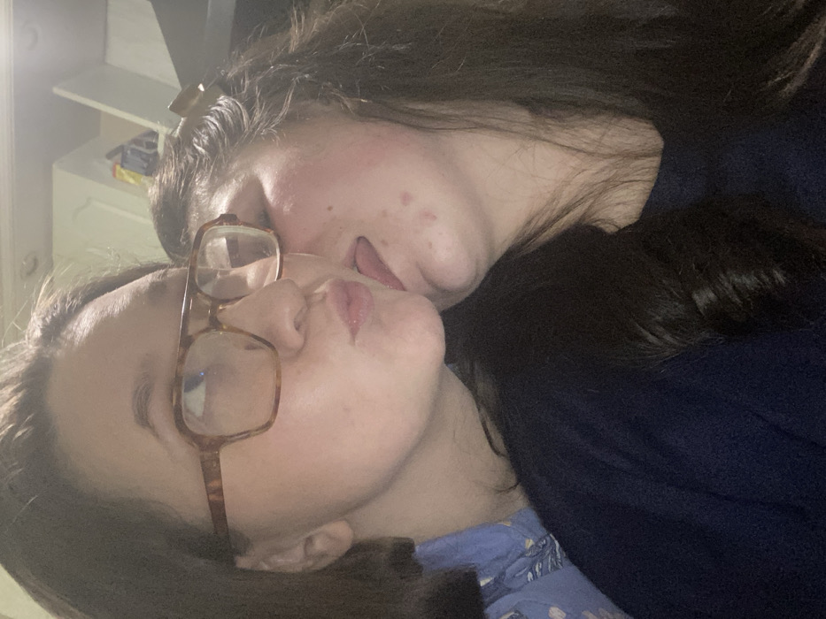 Two women are nestled closely, cheek to cheek. The woman in the foreground wears glasses and a blue patterned top. The other woman is partially obscured, wearing a dark blue top. Both have dark hair. Their tongues are slightly visible, hinting at a playful or intimate moment. The background includes a white dresser with items on top. The image is dimly lit, creating a soft and personal atmosphere.