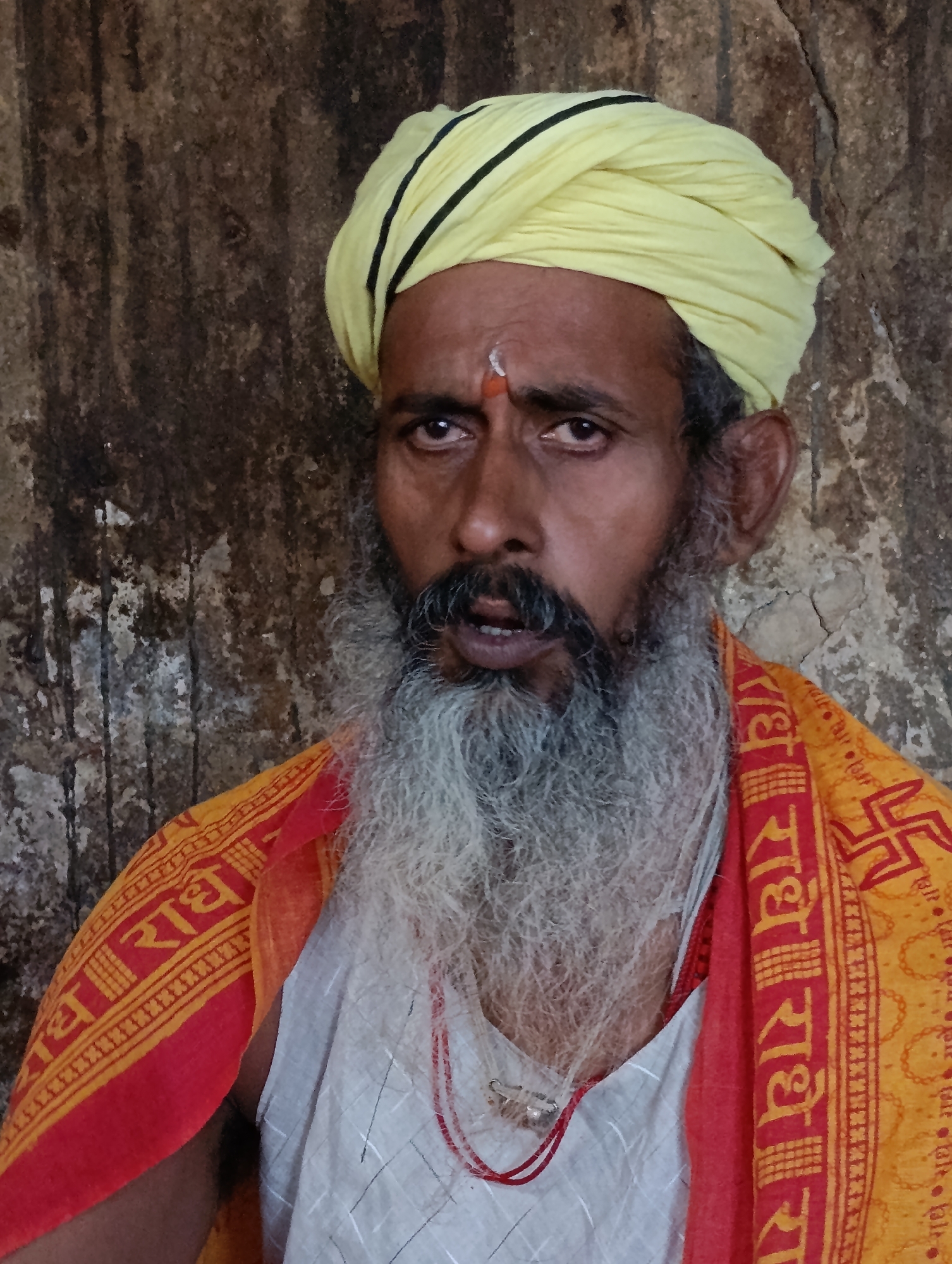 A close-up portrait shows a middle-aged man with a long, gray beard, a yellow turban with black stripes, and a red and yellow sash. He has a red dot on his forehead. The background is a rough, textured wall. The man appears to be in a religious or spiritual setting, possibly in the city of Vashisht, India, known for its temples and ashrams. His expression is solemn and direct, looking towards the viewer.