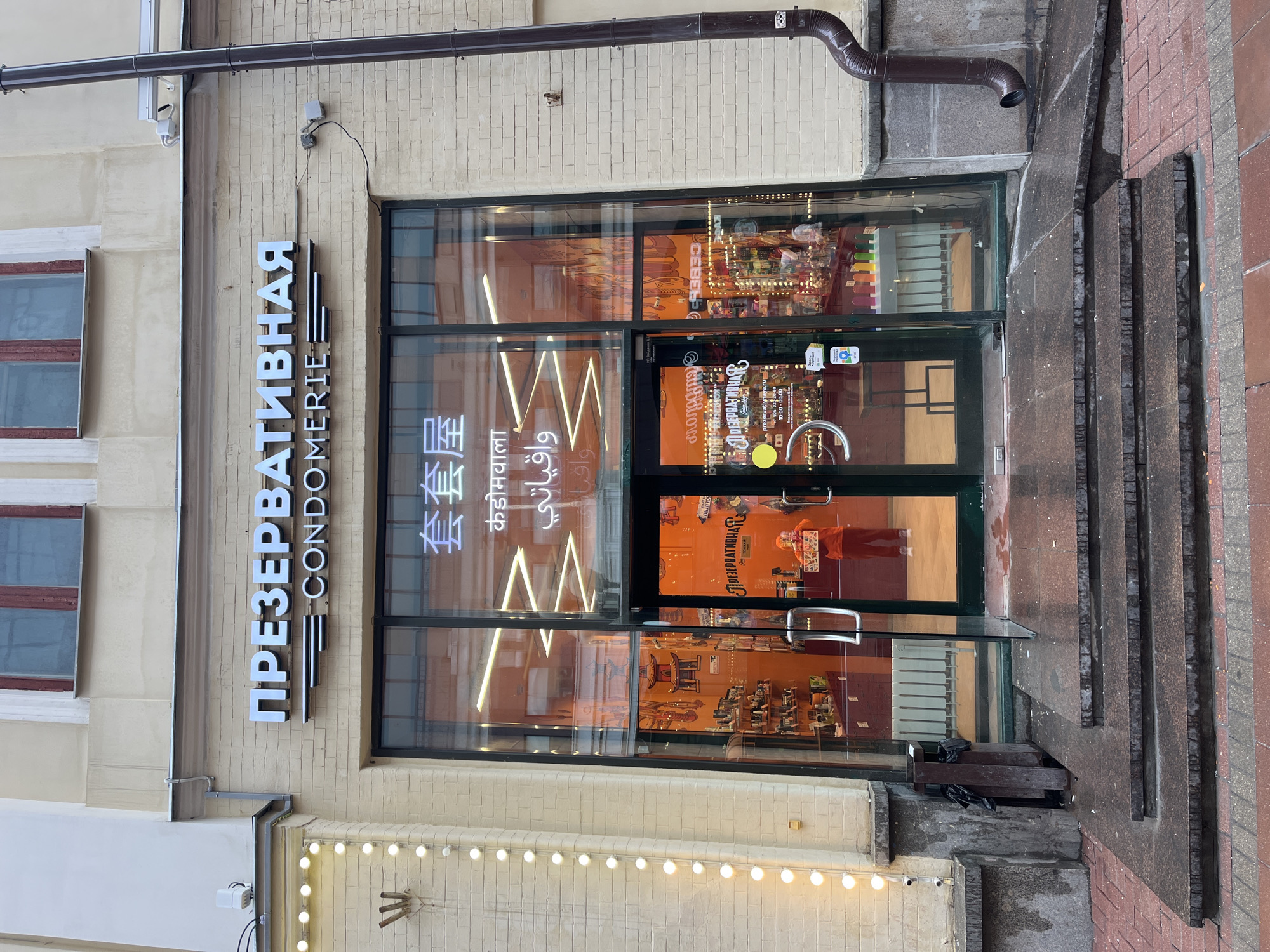 The image depicts the storefront of a business in Moscow, Russia called "Презервативная Condomerie," which translates to "Condom Shop." The name is displayed vertically in white Cyrillic letters on a black plaque affixed to the light yellow brick wall of the building. Below the main title, the name is written in English as "CONDOMERIE." The storefront has a large glass facade with a black frame, and a double glass door with silver handles.
Inside, neon signs written in multiple languages, including Chinese, Hindi, and Arabic are visible. Orange posters and displays are also visible through the glass, containing merchandise. A business hours sticker is affixed to the glass.
The building's exterior has various architectural details, including a brown drainpipe and decorative lights strung along the lower portion of the wall. Steps lead up to the entrance. The sidewalk is made of a combination of stone and brick. A surveillance camera is mounted to the building.