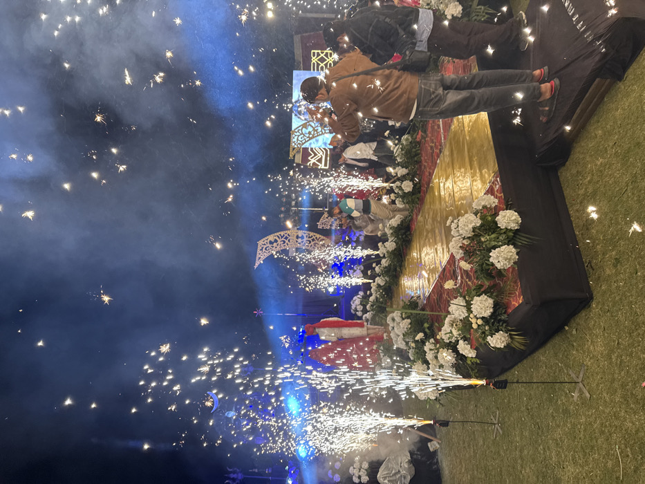 This image captures a vibrant and celebratory outdoor event taking place at night, likely a wedding reception or a grand cultural celebration. The scene is dominated by pyrotechnics and elaborate decorations.
In the foreground, on what appears to be a grassy area, two sparkler machines on thin stands are actively firing, sending brilliant white and gold sparks high into the air. Wisps of smoke from these devices are visible, adding to the dramatic effect.
Central to the image is a decorated stage or platform. The front edge of this stage is adorned with abundant arrangements of lush white flowers, suggesting elegance and festivity. Behind the flowers, the stage has sections of a golden or yellow material, possibly fabric or a backdrop, and a darker, reddish section with a subtle pattern.
On the stage, a person dressed in white with significant red accents, possibly a traditional or ceremonial outfit that includes a red turban and garland, is visible, partly obscured by the intense sparks. Further back on the stage, another individual wearing a light blue and white striped shirt can be seen.
Above the main stage, elaborate, illuminated archways, some with a distinct crescent moon motif, are integrated into the stage design. These arches also appear to be emitting sparks, contributing to the overall spectacular display. Blue spotlights cut through the darkness and the smoke, casting dramatic illumination on various elements of the stage and the people.
In the background, on a slightly elevated area behind the main stage, two men are visible. One man, wearing a brown jacket and dark pants, is holding a smartphone up, seemingly capturing the event with a photo or video. Beside him, another man in a dark jacket observes the spectacle.
The night sky serves as the backdrop, filled with countless scattered sparks, creating a dazzling, almost starry effect across the upper portion of the image. The air is diffused with smoke and the glow of the blue lights and sparklers. The overall atmosphere is festive, energetic, and visually stunning, characteristic of a significant celebration. No discernible text is visible in the image.