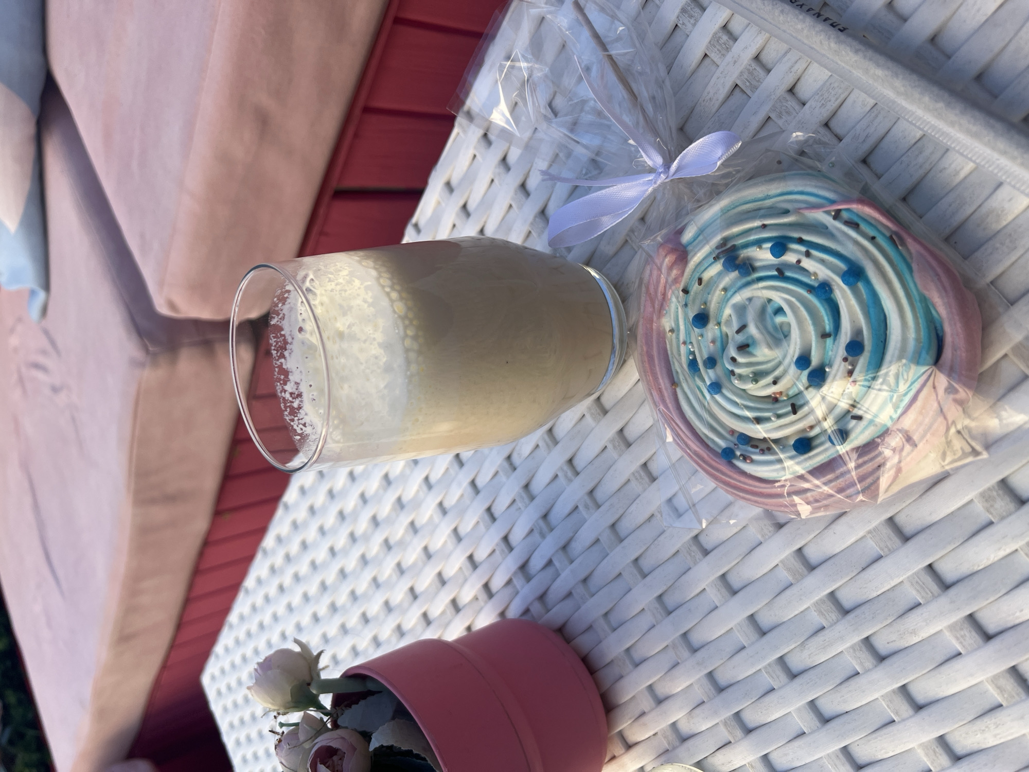 A high-angle, natural light photograph depicts a still life arrangement on a white, woven wicker table or surface. A glass filled with a light brown beverage with a foamy top sits on the table. To the right, a large, swirled meringue lollipop, wrapped in clear plastic and tied with a white ribbon, is also placed on the table. In the lower left corner, a small pink flower pot with miniature white and pink roses adds a touch of floral decoration. The background includes a glimpse of a peach-colored cushioned seat or bench, with a red-toned wooden structure behind it.
Although the style of the images suggests an outdoor setting suitable for Plovdiv, Bulgaria, there is not any way to verify the exact location, thus no information can be determined about the location of Plovdiv, Bulgaria. There is a lack of sufficient context in the image.