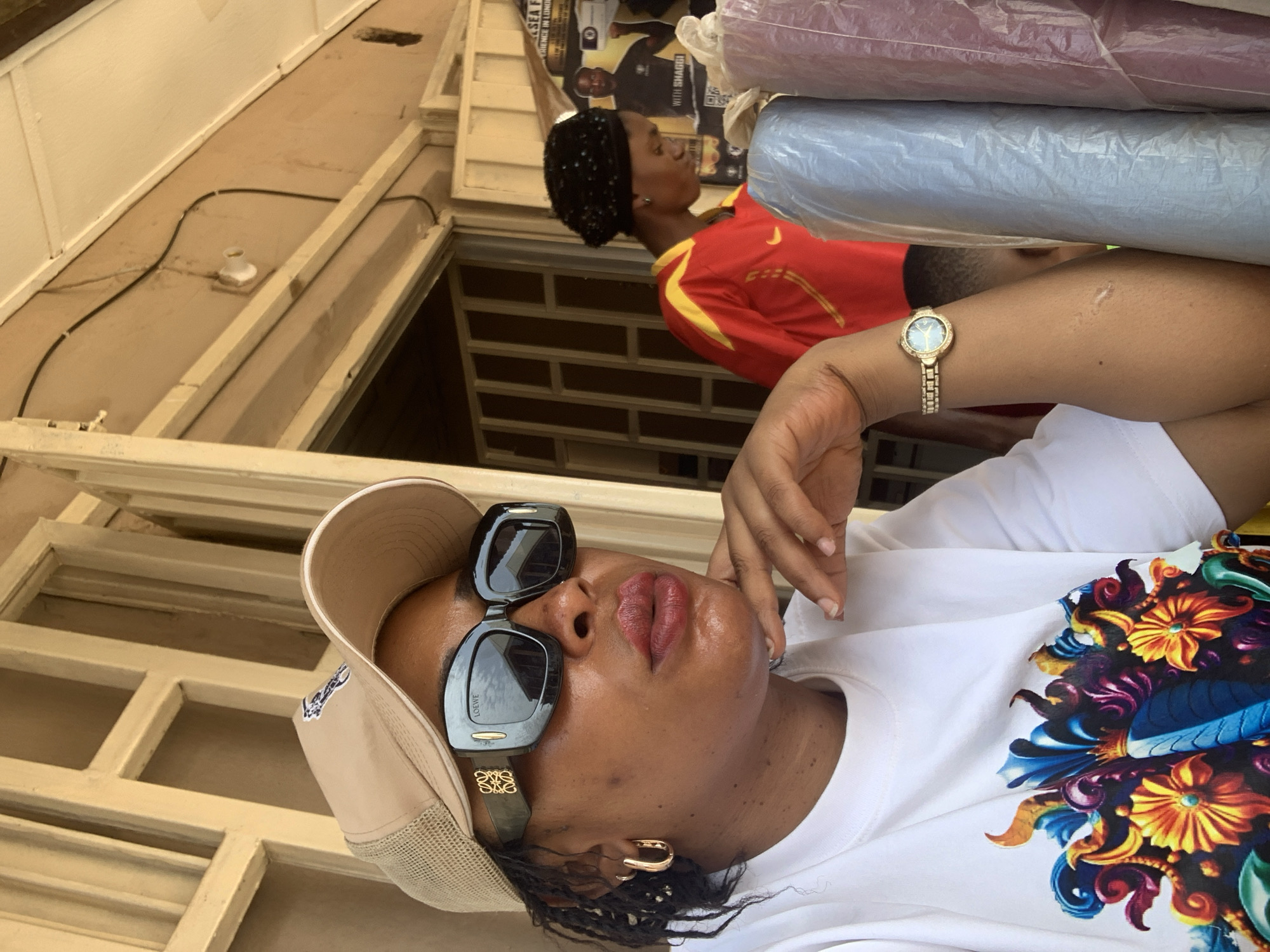 A medium shot captures a woman in Sabon Tasha, Nigeria, wearing a beige baseball cap, sunglasses, a white t-shirt with a multicolored floral design, and a gold watch. The woman appears to be resting her chin on her hand. Behind her, another person wears a red and yellow t-shirt and carries stacked fabrics or materials. A poster is affixed to the wall behind the second person. Wooden window frames and a building facade surround them.