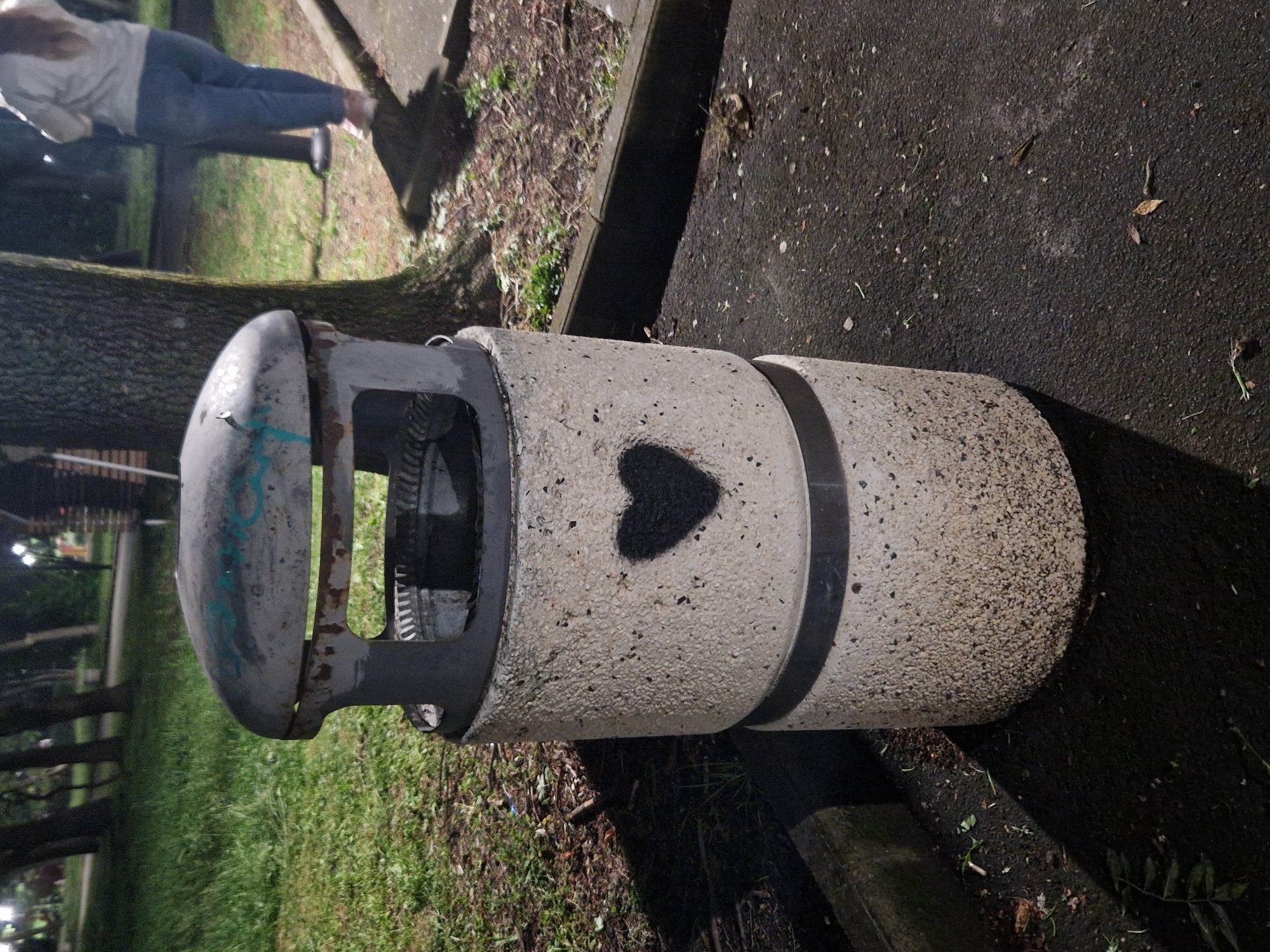 The image, rotated 90 degrees clockwise, captures an outdoor scene at what appears to be dusk or night, illuminated by artificial lighting. In the foreground, a sturdy cylindrical trash bin is the main subject. It is constructed from light-colored, speckled concrete, and features a distinct black heart shape stenciled on its side. The top section of the bin is made of darker gray metal, showing signs of weathering and rust, with faint turquoise or light blue graffiti.
Behind the trash bin, a thick tree trunk is visible. The ground cover consists of green grass interspersed with dry leaves and debris, alongside a dark asphalt surface that has a fine speckled texture. A concrete curb runs along the edge of the asphalt.
In the upper left background, a person is partially visible, seen from behind. They are wearing a light-colored long-sleeved top and blue jeans, and appear to be walking away from the viewer. Their features are not discernible. In the far background, blurred lights suggest distant urban illumination.
The scene depicts a common urban or park environment. No specific event is taking place beyond casual movement in a public space. There is no discernible text in the image.