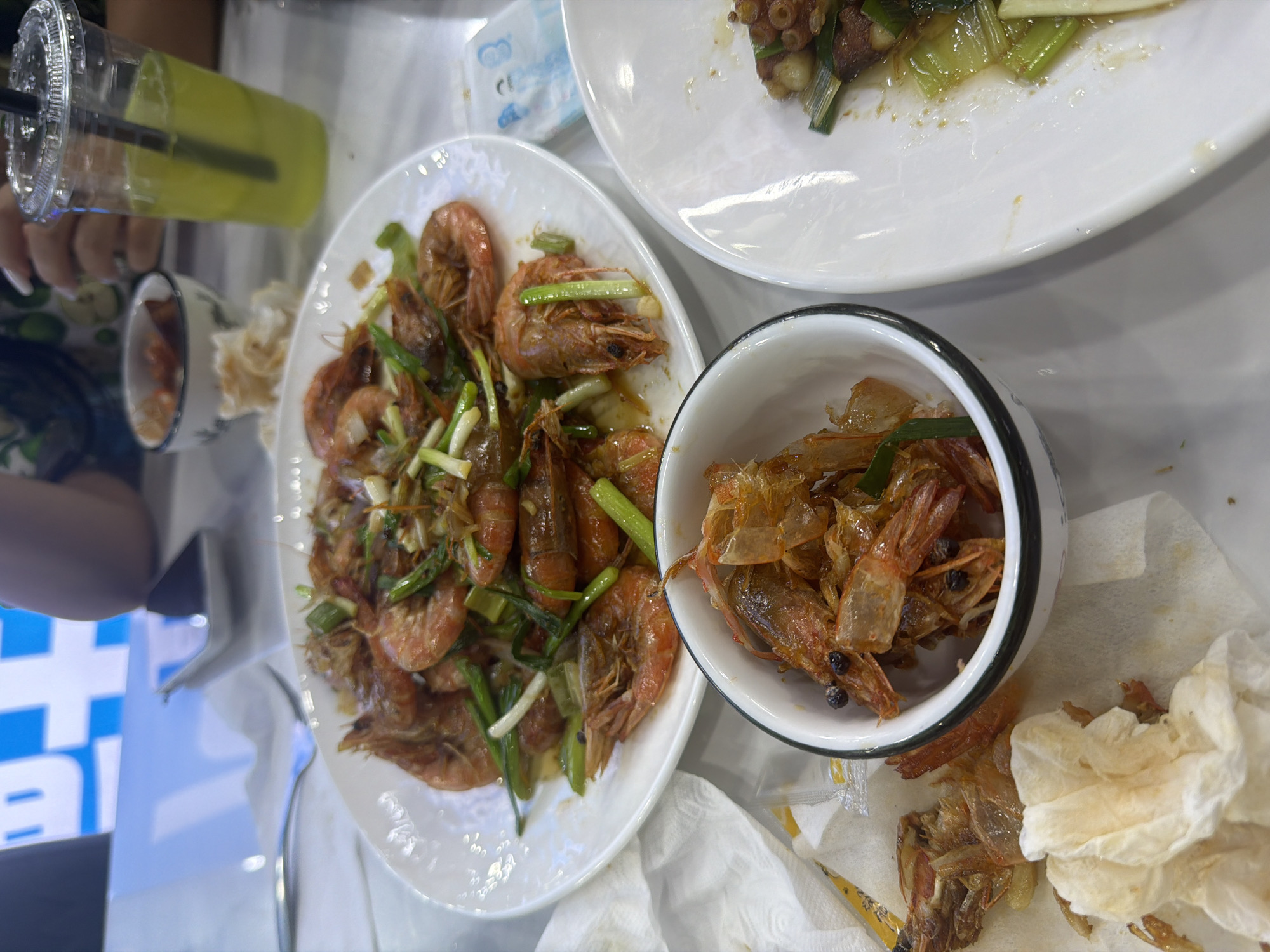 This image captures a close-up view of a table laden with food and drinks, suggesting a meal is in progress. The primary subjects are plates of cooked shrimp, some whole and garnished with green onions, and a smaller bowl containing what appears to be discarded shrimp shells and some whole shrimp. There's also a white plate with some cooked food, possibly octopus or another seafood dish, with green onions. A clear plastic cup filled with a yellow-green beverage and a black straw is visible, held by a person whose hands and fingernails are partially in frame. The setting appears to be indoors, likely a restaurant, with a white tablecloth and some background elements that hint at a busy environment, including a blue and white graphic sign. The lighting suggests it could be daytime or evening. The overall mood seems casual and perhaps focused on the enjoyment of the meal, with the abundance of food and drink. There is no discernible text in the image.
