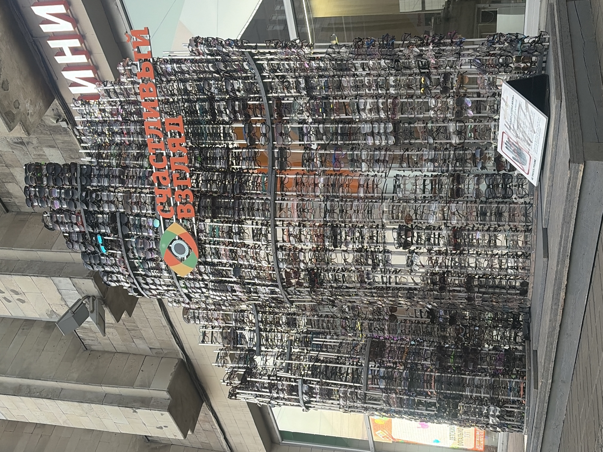 The image shows a large, multi-tiered display structure made of numerous eyeglasses, resembling a tower or sculpture. The structure is located outdoors, in front of a building with a large window displaying signage. The building appears to be a retail establishment, possibly an optical shop, given the display of eyeglasses. The signage above the window reads "ИНИ" in large white letters on a red background. Below this, in orange lettering on the eyeglasses structure, are the words "СЧАСТЛИВЫЙ ВЗГЛЯД", which translates to "Happy Look" or "Happy Gaze". There is also a circular logo with an eye design. In the foreground, on a wooden platform, is a white sign with text and an illustration of a hand. The overall setting suggests an urban environment, likely a commercial street in Saint Petersburg, Russia, as indicated by the building architecture and the Cyrillic lettering. There are no people visible in the image. The lighting suggests it is daytime, possibly overcast.