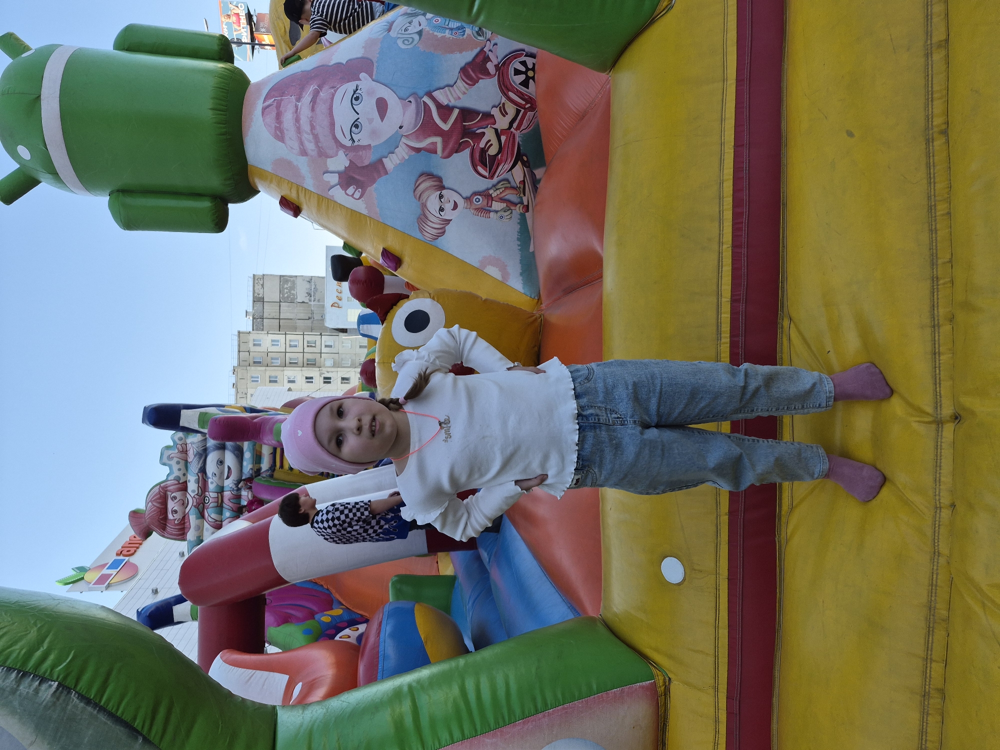 A young girl with a pink hat and long braids is lying on her back on a yellow inflatable surface with a red stripe, wearing a white long-sleeved shirt and blue jeans with pink socks. She is looking at the camera. The inflatable structure is part of a larger inflatable play area with cartoon characters and shapes, including a green Android figure. A building is visible in the background. The location is Tobolsk, Russia, as indicated by the Cyrillic text on a sign.
