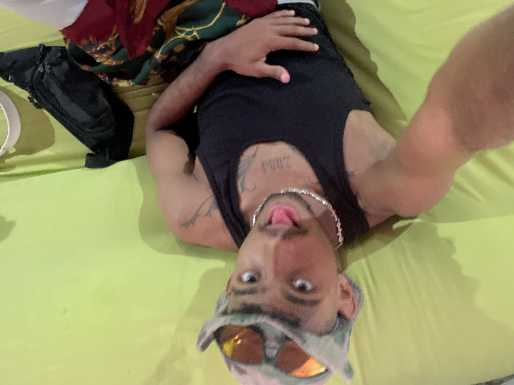 An individual wearing a camouflage hat with orange-tinted sunglasses is lying on a bright green surface, possibly a bed or couch. The person is wearing a black tank top and has a silver chain necklace. A tattoo is visible on their chest, with the number "2004" prominently displayed. The individual's tongue is sticking out. To the upper right of the individual, a black fanny pack is visible, along with a colorful fabric, possibly a blanket or clothing. The background is a solid green color. This scene could be located in a bedroom or living area in the city of La Asuncion, Venezuela.