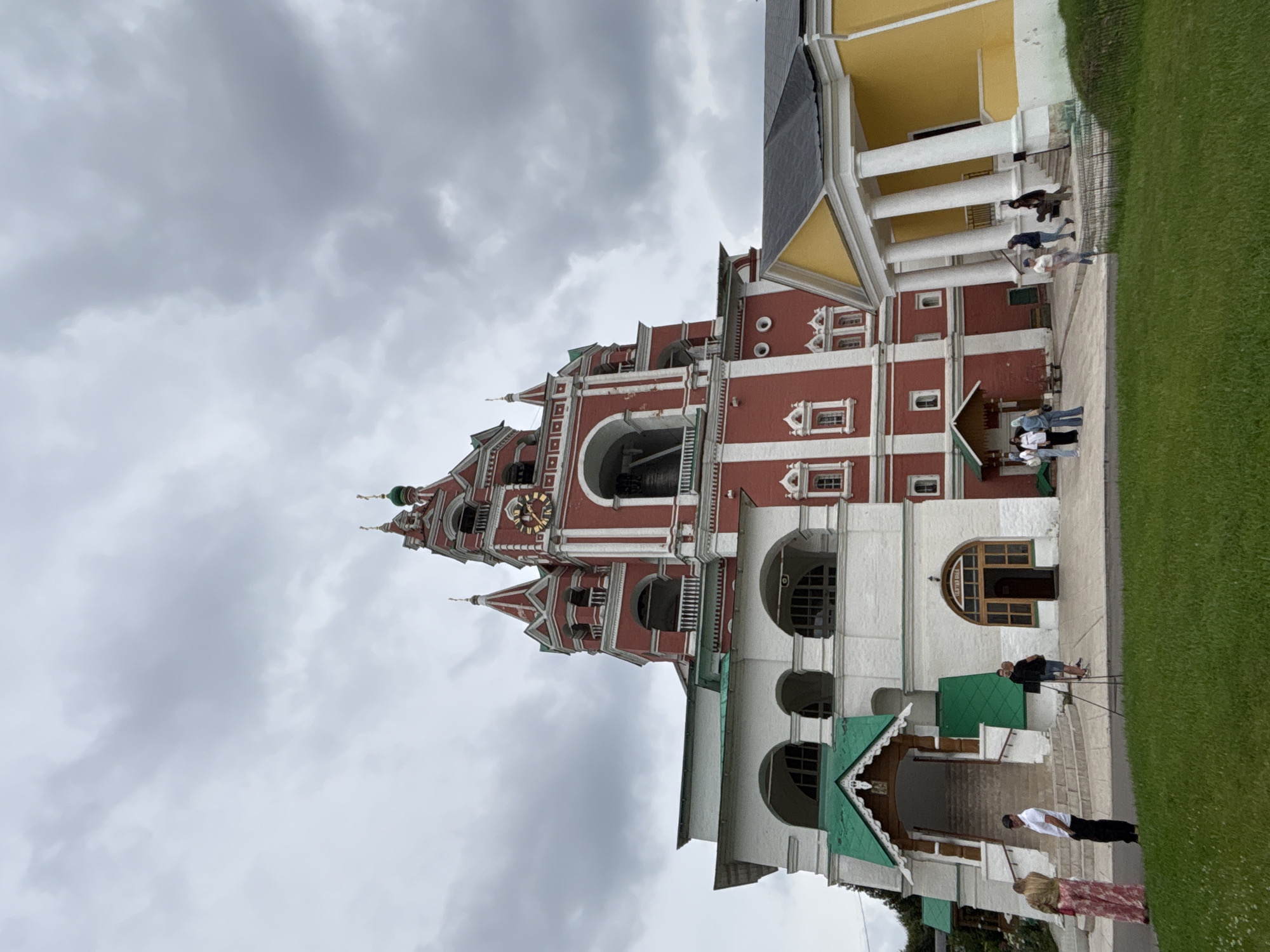 This is a low-angle, medium shot of the Church of the Nativity of the Theotokos at the Savvino-Storozhevsky Monastery in Zvenigorod, Russia. The church's red bell tower with white trim and green domed spires dominates the frame, partially obscured by an overcast sky. To the left of the bell tower is a white, two-story section of the monastery with green roofs. To the right, a yellow building with white columns and a dark roof extends into the frame. Several people are visible in the scene, appearing as small figures against the backdrop of the large structures. A woman in a long, patterned dress stands on the left, near a man in a white shirt and black pants. Further to the right, a group of people are gathered near the entrance of the yellow building. The foreground consists of a well-maintained green lawn.