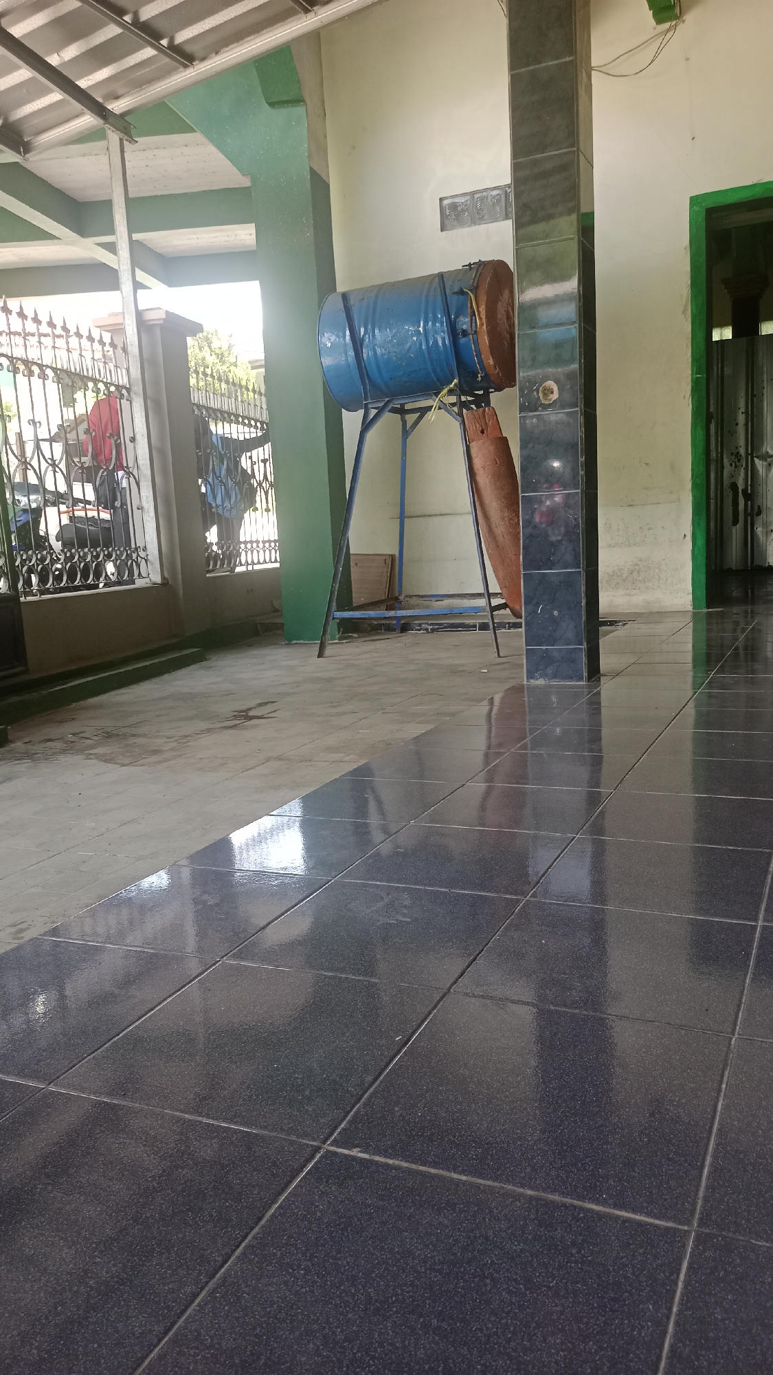 The image depicts a scene in Karyamandala, Indonesia, showing the exterior of a building with a tiled floor. In the foreground, dark, glossy square tiles reflect the ambient light. A column covered in black tiles is visible on the right side of the frame. Further back, a blue barrel, possibly a drum, is mounted on a metal stand. A reddish-brown object, also barrel-shaped, hangs below the blue drum. The background reveals a metal fence with decorative patterns, a glimpse of a motorcycle, and a green-painted pillar. The walls of the building are painted in muted tones, and a doorway with a green frame is visible to the far right. The scene appears to be outdoors, under a covered structure with a visible metal roof framing.
