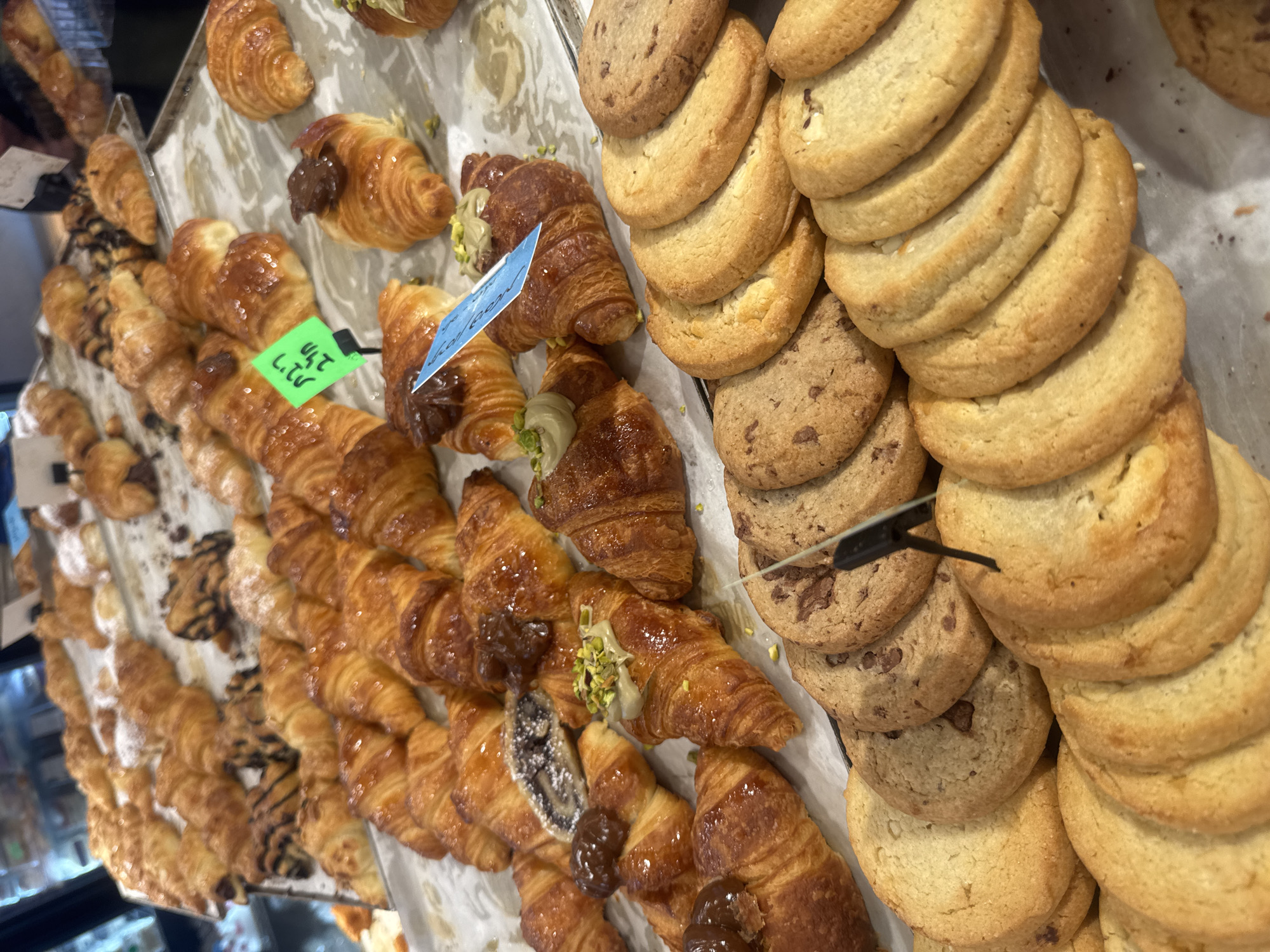 An elevated, angled shot displays an assortment of pastries in a bakery, likely in Ramat Gan, Israel, given the Hebrew writing on a green price tag. The foreground is dominated by stacks of pale yellow cookies with visible crackled surfaces. Adjacent to the cookies are rows of golden-brown croissants, some filled with chocolate or topped with green pistachio crumbles. The croissants are of varying sizes and styles. Price tags are visible among the pastries. The pastries are displayed on a metal tray. The overall lighting is bright.