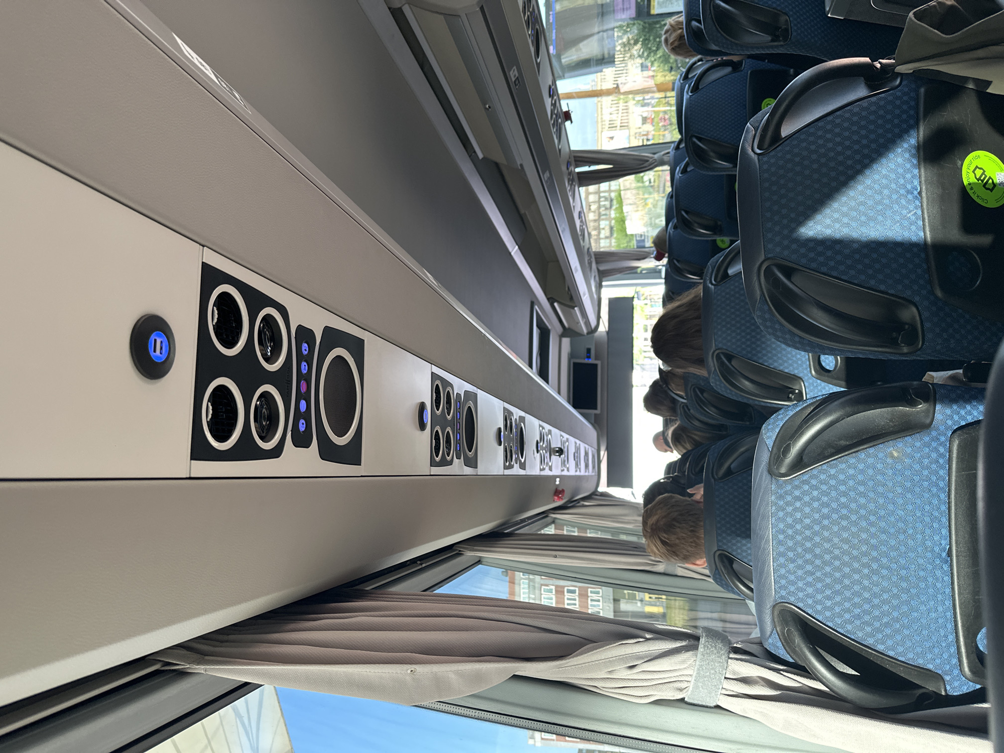 The image shows the interior of a bus with several passengers seated. The camera is positioned looking forward, towards the front of the bus, from the perspective of a passenger. Blue upholstered seats with black armrests are visible. The ceiling of the bus has overhead compartments and what appear to be air vents and USB charging ports. Curtains are drawn to the left side of the image, revealing a glimpse of buildings and the sky outside. One passenger in the foreground has their back to the camera, with brown hair. Other passengers are visible further ahead, also with their backs to the camera. The overall scene depicts a typical bus journey. There is no indication of the city of Utrecht, Netherlands, in the image itself.