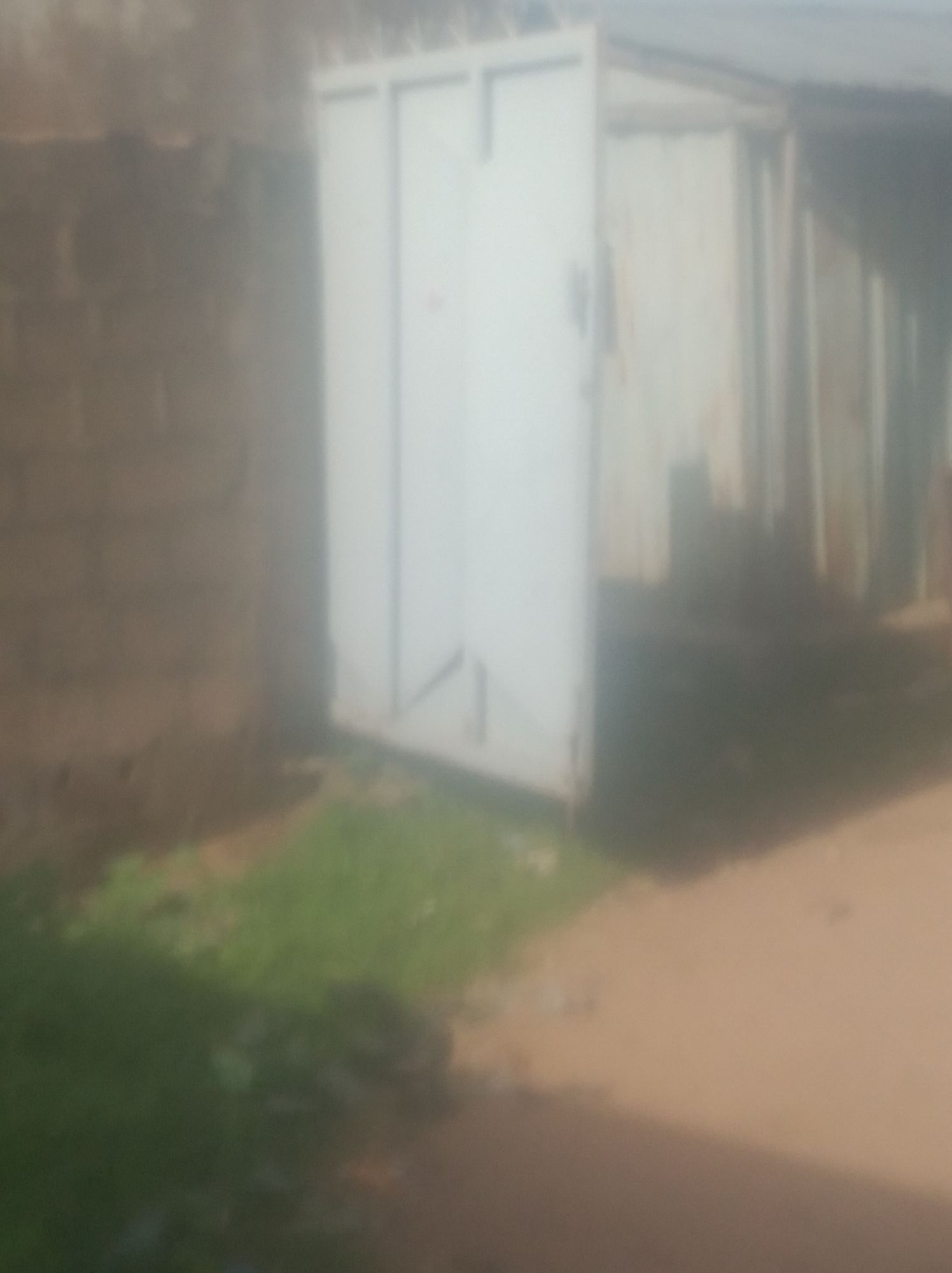 The image shows a blurred view of a building exterior in Jalingo, Nigeria. On the left, there is a section of a brick wall. To the right of the brick wall, there is a large, open, white metal gate or door, which appears to be made of corrugated metal panels. The gate is partially ajar, revealing a dark interior or the back of the structure. The ground in front of the gate is covered with patchy green grass and dirt. In the background, more of the building's structure is visible, with vertical lines suggesting paneling or slats, and a roofline is suggested at the top. The overall image is out of focus, making it difficult to discern finer details.