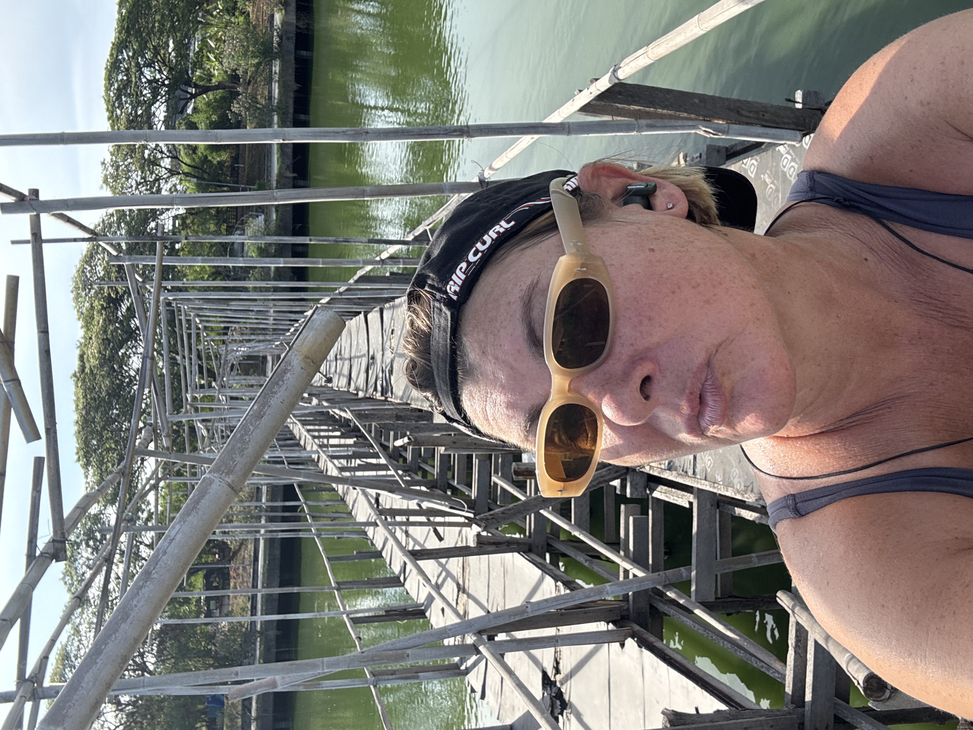 A woman wearing sunglasses and a "Rip Curl" hat is standing on a bamboo bridge in Baan Chaweng, Thailand. The bridge appears to be under construction or repair, with bamboo scaffolding. Green water is visible beneath the bridge. The woman is facing the camera, suggesting a selfie. Trees are visible in the background.