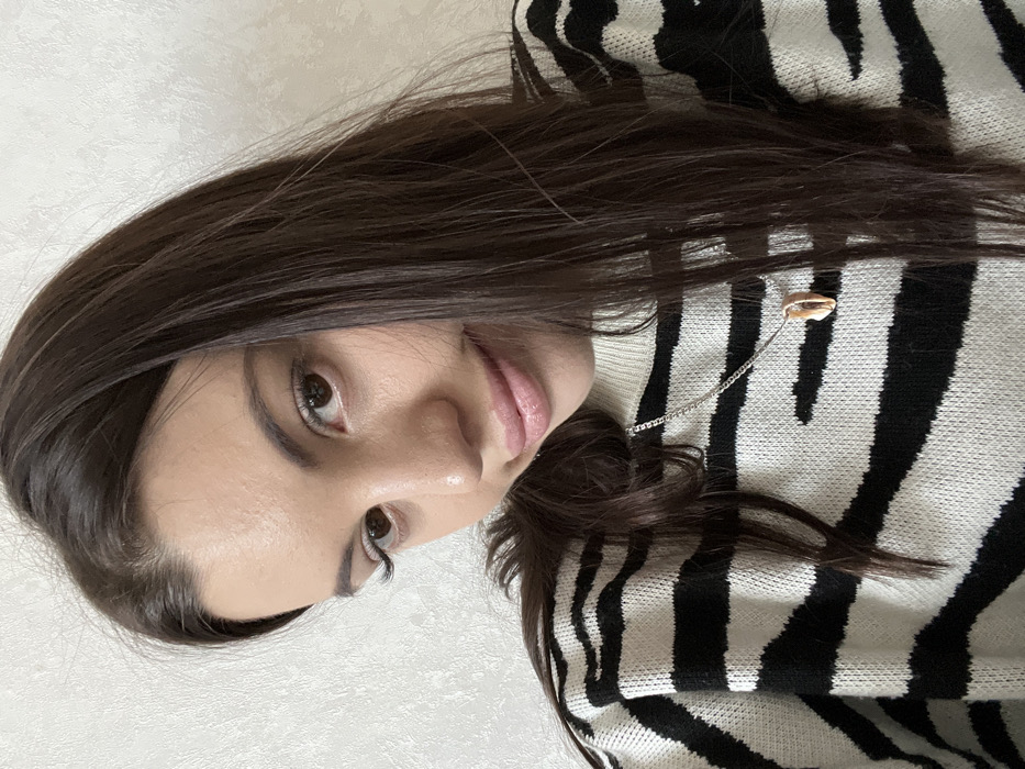 A woman with long, dark hair poses in a selfie-style shot. She wears a black and white zebra-striped sweater and a delicate silver chain necklace with a seashell pendant. Her skin is fair, and she has light brown eyes. The image is slightly tilted, showcasing her face from a close-up angle. A subtle smile plays on her lips, adding a gentle touch to the portrait. The background is plain and light-colored.