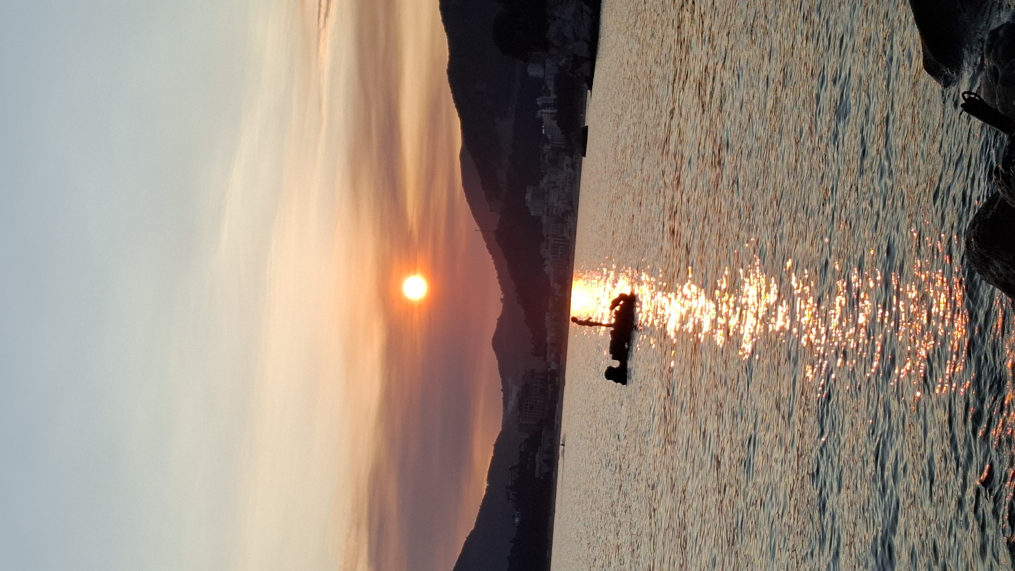 A vertical, low-angle, full shot captures a sunset over the Adriatic Sea in Budva, Montenegro. The sun, a bright orb, is positioned in the center of the frame, slightly to the left, casting a warm orange glow across the sky. Clouds, tinged with pink and orange, stretch across the upper portion of the image, transitioning to a pale blue at the top.
In the mid-ground, a silhouette of a person stands on a dark, irregularly shaped object in the water, likely a rock formation. The person appears to be standing or possibly engaged in an activity such as fishing or paddleboarding, though their exact action is indistinguishable due to the silhouette.
The sea is calm, with gentle ripples reflecting the sunlight in a shimmering path that extends from the horizon towards the foreground. The water is a deep blue-green, catching the warm hues of the sunset.
In the background, the coastline of Budva is visible, characterized by rolling hills covered in lush vegetation and dotted with buildings that form a town or city. The structures are mostly dark silhouettes against the horizon, with faint lights suggesting an inhabited area. The overall atmosphere is serene and picturesque, highlighting the natural beauty of the Montenegrin coast at dusk.