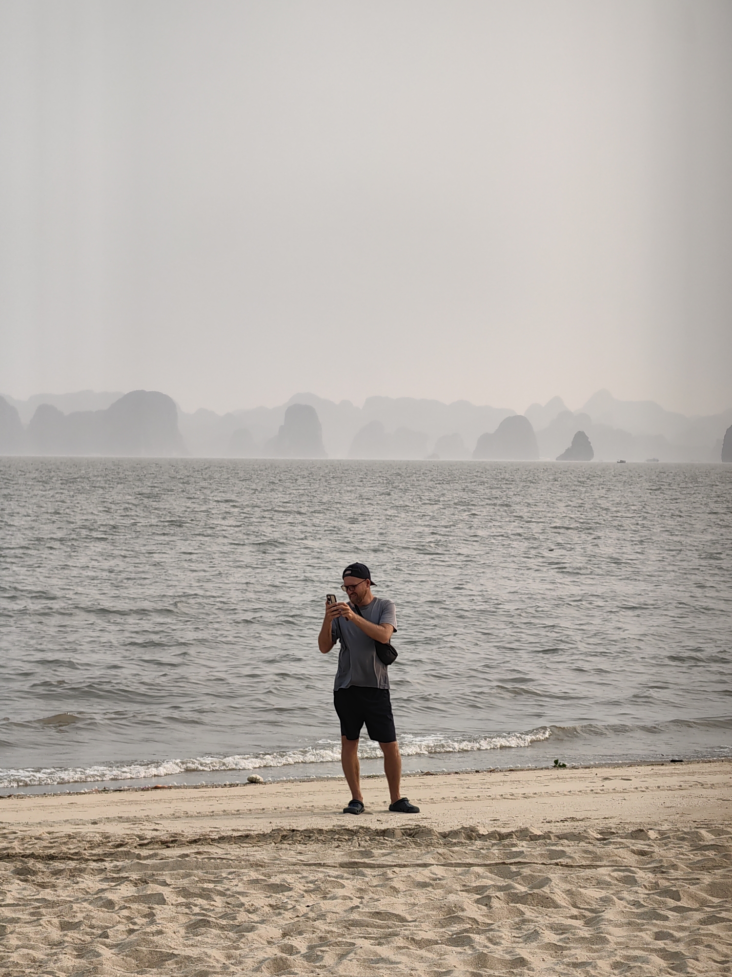 The image depicts a man standing on a sandy beach in Bai Chay Ward, Vietnam. He is holding a smartphone and appears to be taking a photograph. The sea stretches out behind him, with hazy islands visible in the distance. The sky is overcast. The sand is textured with footprints and other marks. The man is wearing a dark baseball cap, glasses, a gray t-shirt, dark shorts, and dark sandals. He has a black bag slung over his shoulder. The water's edge shows small waves lapping onto the shore.