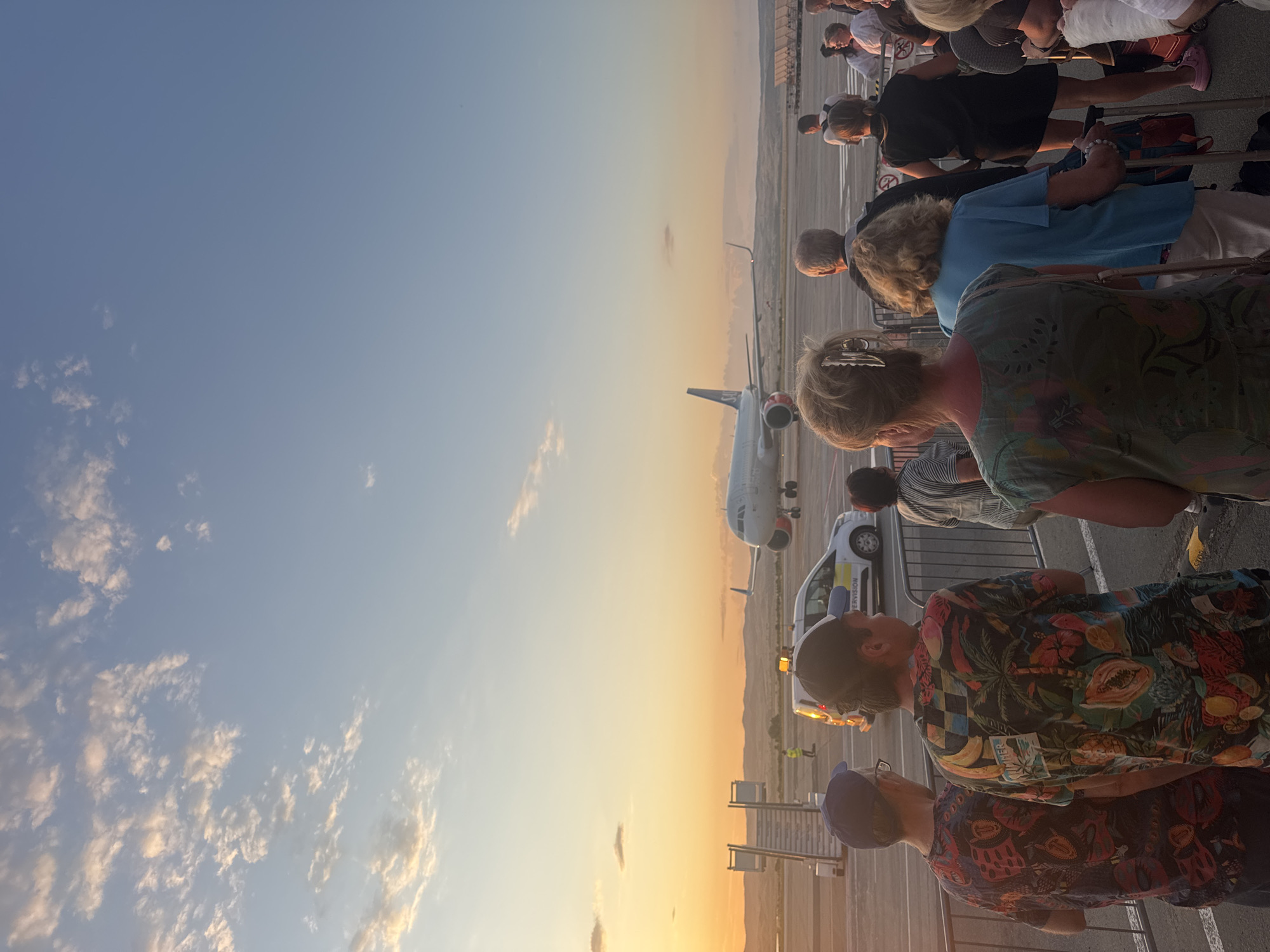 A group of people stand in line on an airport tarmac at sunset. The sky is a gradient of blue and orange, with scattered clouds. An airplane is visible in the background, taxiing on the runway. In the foreground, a white airport vehicle with a flashing yellow light is parked. The people are facing away from the camera, towards the plane. The overall scene depicts a typical airport boarding or disembarking situation at dusk. The location is Burgas, Bulgaria.