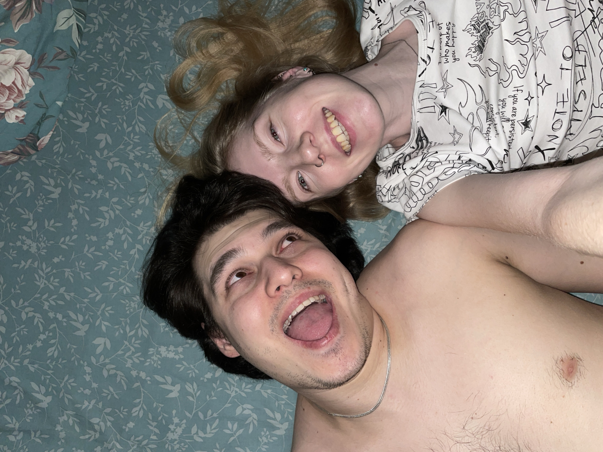 Two individuals, a man and a woman, are lying down side-by-side, facing upwards and slightly to the right, creating a casual and intimate scene. Their heads are positioned towards the center of the frame, with their bodies extending outwards.
Both subjects display expressions of joy and playful excitement. The man, with dark hair and light skin, has his mouth wide open, as if in mid-laugh or exclaiming, and looks upward. He is shirtless, revealing his chest and a silver chain around his neck, and has visible facial stubble. The woman, with long blonde hair spread around her, mirrors his upward gaze with a broad, happy smile. She has multiple ear piercings and a nose ring, and is wearing a white t-shirt adorned with black graphic designs and text.
The setting appears to be an indoor space, likely a bedroom, indicated by the patterned bedding beneath them. The left side of the bedding features a blue-green fabric with large, faded floral patterns in pink and white, while the rest displays a lighter blue-green pattern with delicate white leaves and bird motifs.
The lighting suggests an indoor environment, possibly at night or in low ambient light conditions. The overall emotion conveyed is one of happiness and lightheartedness. Visible text on the woman's t-shirt includes phrases such as "who makes you happy," partially visible text that reads "If you are persistent you will keep it consistent," and "NOTE," interspersed with abstract black drawings like stars and flames. This candid moment takes place in Izhevsk, Russia.