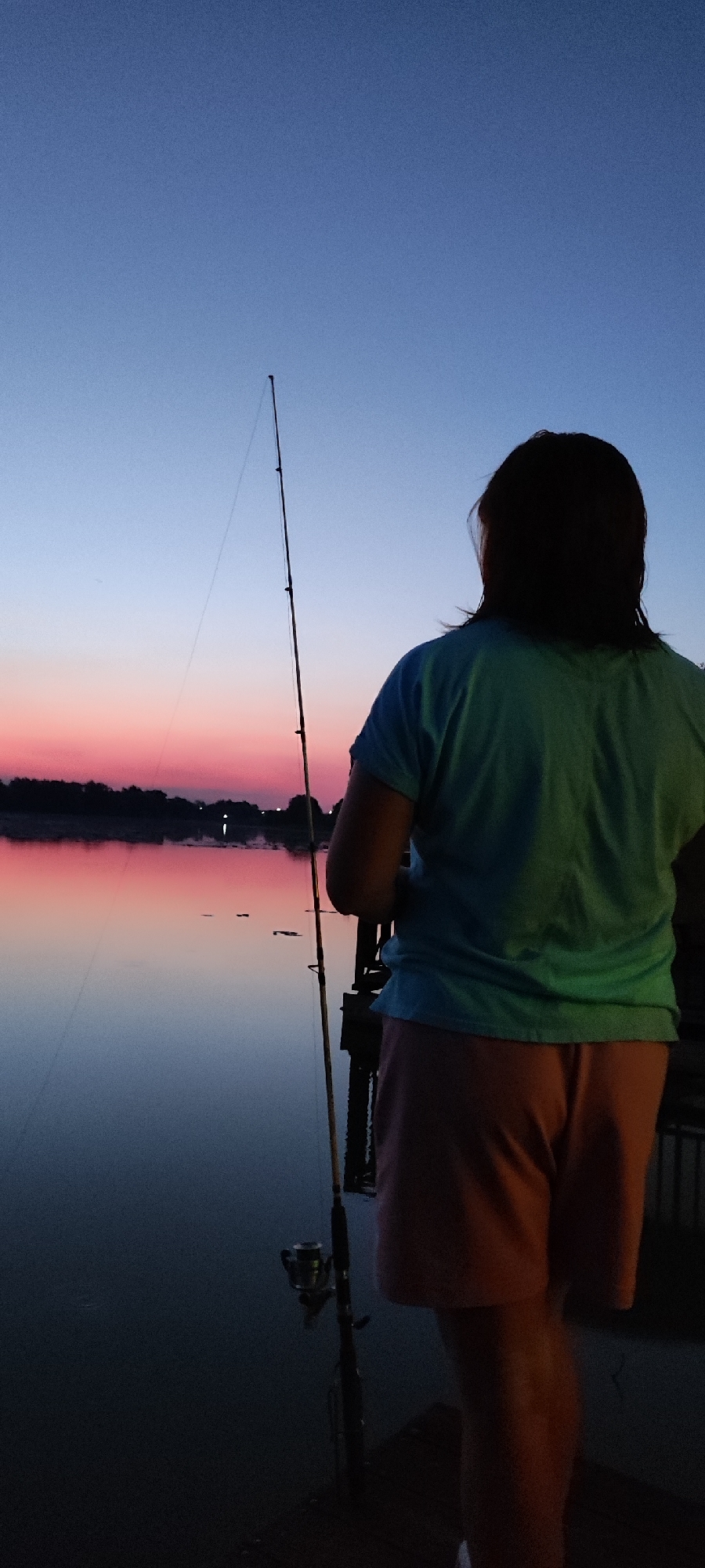 A person stands on a wooden pier at dusk, fishing. The sky transitions from a deep blue at the top to a soft pink and orange gradient near the horizon, reflecting on the calm water. The person, viewed from behind, wears a turquoise t-shirt and peach-colored shorts. They are holding a fishing rod, which is angled upwards and to the left. A fishing reel is visible near the base of the rod. Silhouetted trees line the far bank of the water, with a few faint lights visible in the distance, suggesting a populated area. The overall mood is tranquil and serene, capturing the quiet beauty of a sunset by the water. The time of day is clearly evening, as indicated by the sunset colors and the dimming light. There is no visible text in the image.