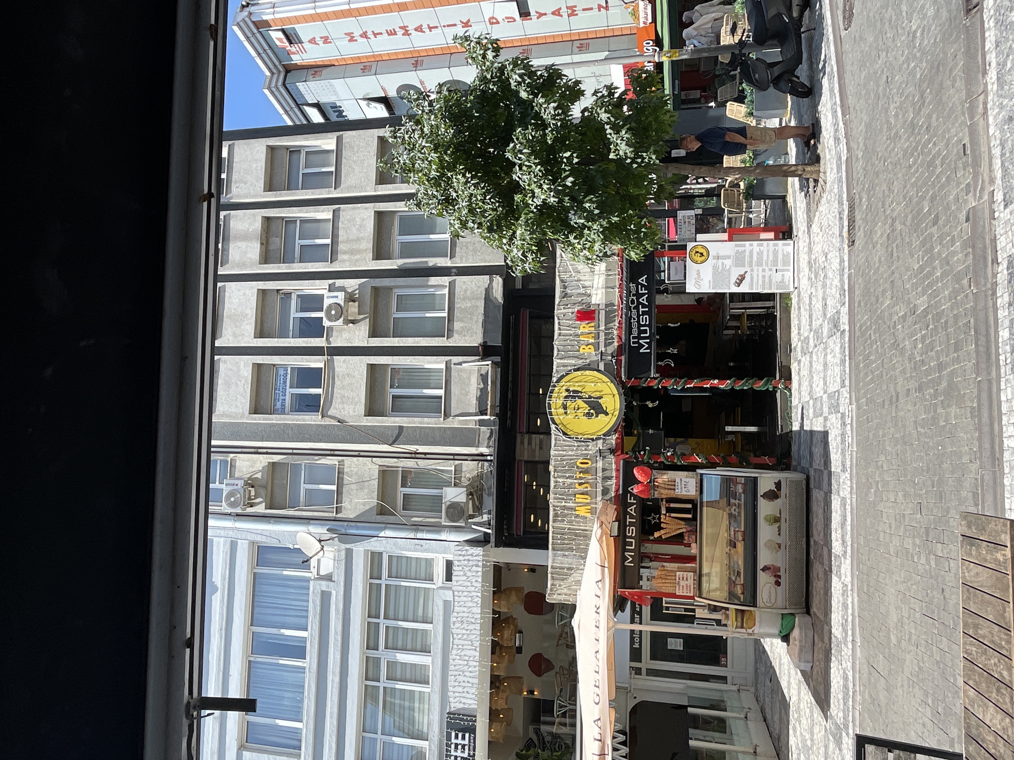 The image shows a street scene in Kadıköy, Turkey, likely on a sunny day, with a focus on commercial establishments. In the foreground, a cobblestone street is visible, with a wooden table in the lower left corner. To the right of the table, an ice cream display case is filled with various flavors of ice cream. Ice cream cones are also displayed above the case. A sign above the ice cream stand reads "Mustafa".
Behind the ice cream stand, a building with multiple floors is visible, with shops and restaurants on the ground level. One establishment has a prominent sign that says "MasterChef Mustafa". Another sign reads "MUSTAFA BAR". A tree with green leaves stands to the right of the building, providing shade.
A man in a blue shirt and khaki shorts is standing near the tree, looking up. Further to the right, a black scooter is parked. In the background, other buildings with windows and air conditioning units are visible. Signs for businesses are present on the buildings, including one with Turkish text that appears to be related to mathematics. The sky is clear and blue. The overall impression is of a lively street in a Turkish city, likely a popular area for shopping and dining.