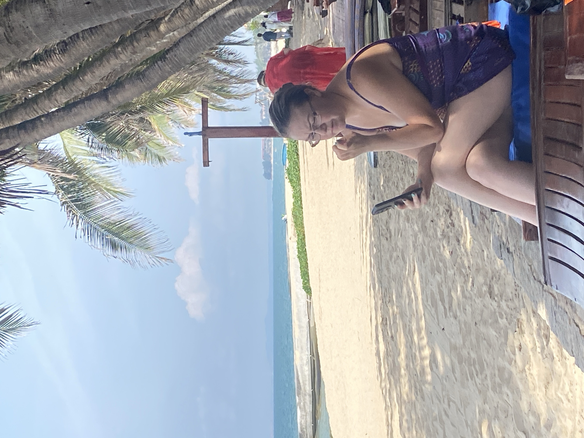 A woman wearing a purple patterned dress and glasses sits on a beach chair on a sandy beach. She is holding a cell phone in her left hand and her right hand is near her face. In the background, the ocean is visible under a bright blue sky with a few clouds. Palm trees cast shadows on the sand, and other beachgoers are walking in the distance. The scene appears to be at a resort in Phu Thuy Ward, Vietnam.