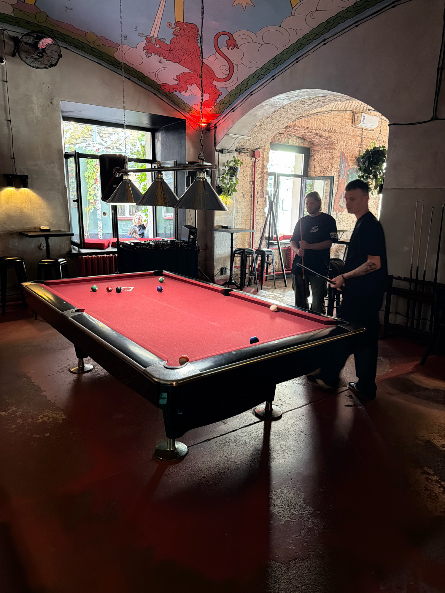 Two men are playing pool in a dimly lit room with a red pool table. The room has a painted ceiling with a red lion and clouds. There is a large window behind the pool table, showing some greenery outside. A DJ setup is visible near the window. The floor is a dark red color and appears to be concrete or asphalt. The overall atmosphere suggests a casual bar or club setting. The location is in Moscow, Russia.