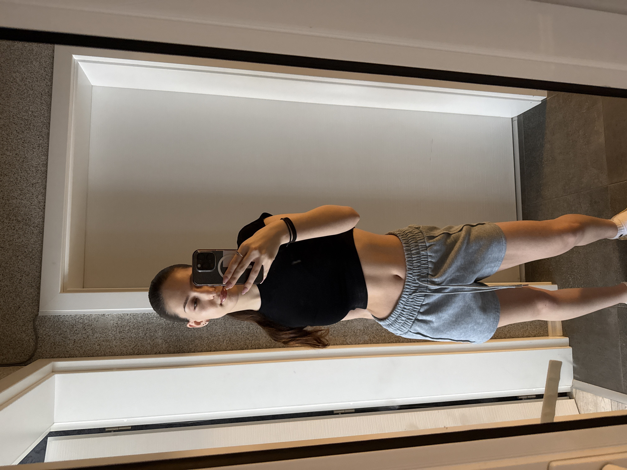 A young woman is taking a selfie in a mirror. She is wearing a black crop top and grey shorts. She is holding a smartphone in her right hand, partially obscuring her face. The background consists of a white door frame, with different colored flooring on either side of the frame. The photo was likely taken indoors. There is no information to indicate that the location is in Sofia, Bulgaria.
