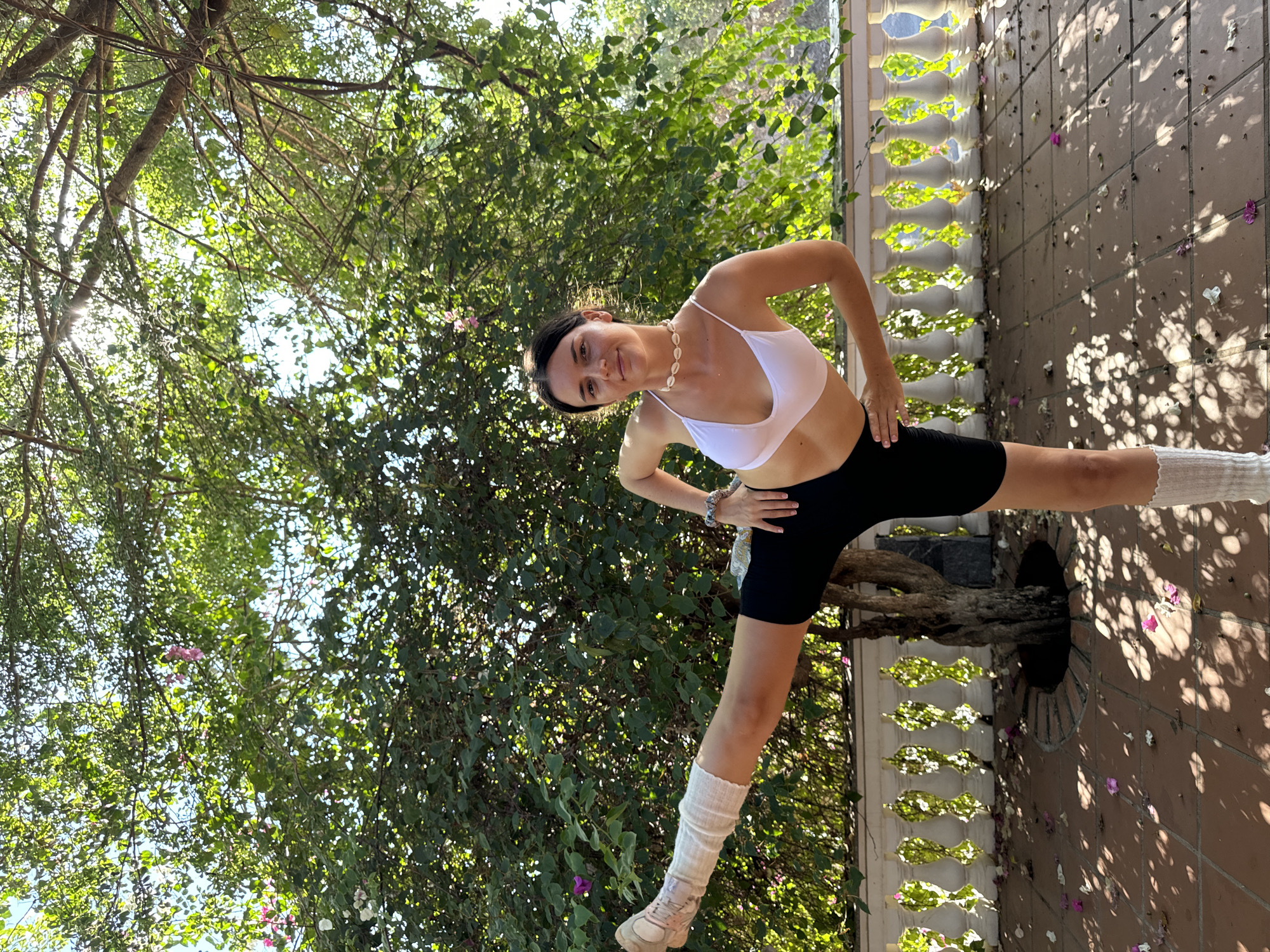 A woman is captured in a full-length shot, positioned on a tiled terrace. She is clad in a white sports bra and black athletic shorts, complemented by high socks and athletic shoes. The woman is performing a yoga stretch, one leg extended to the side, while her hands rest on her hips.
The terrace features a decorative white railing, and beyond it, lush green foliage is visible, filling the background. The environment is sun-drenched, with shadows of leaves dappling the terrace. The scene is located in Mũi Né, Vietnam.