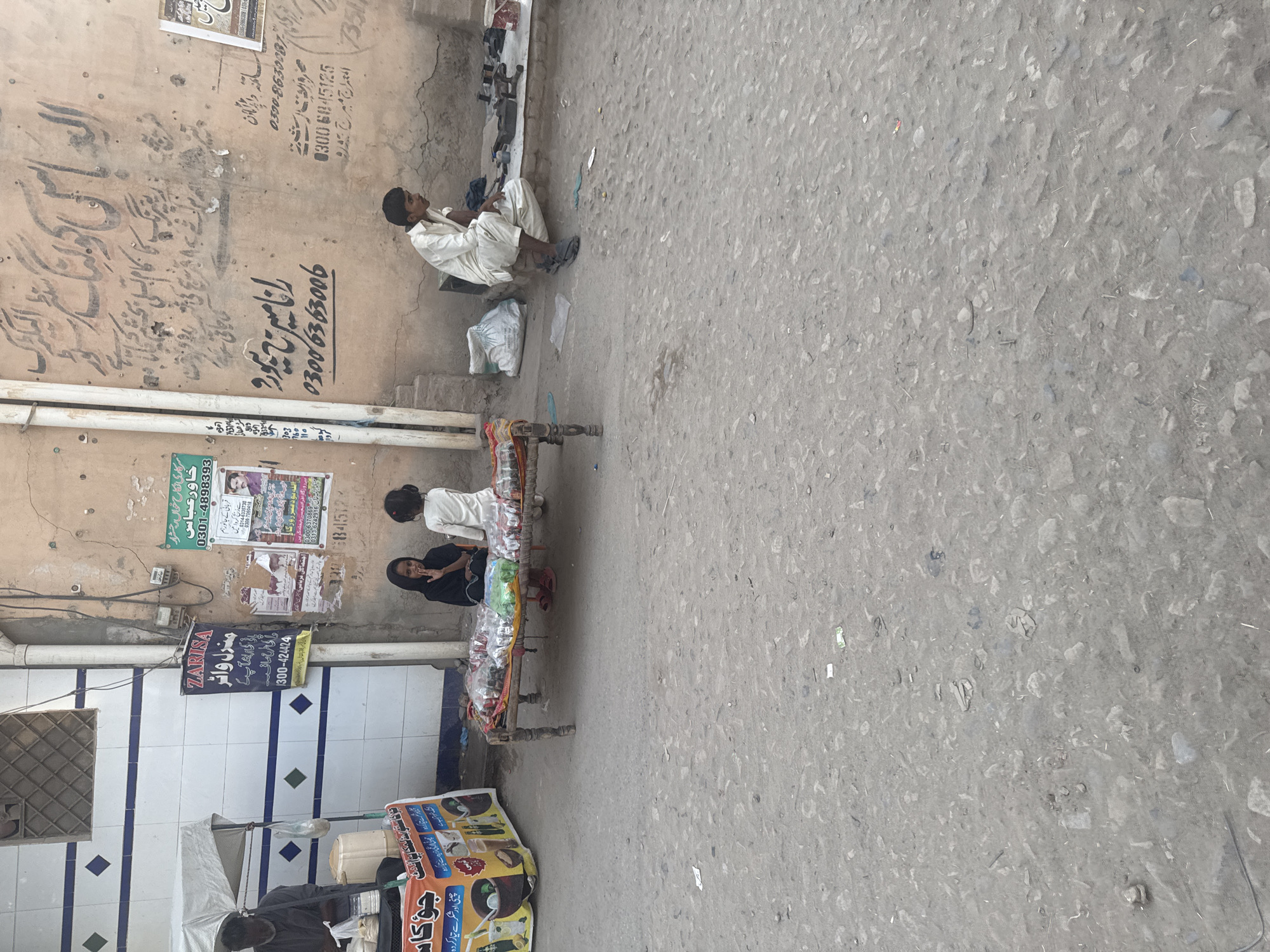 The image depicts an outdoor street scene in Unknown, Unknown, characterized by a dusty, unpaved road and aged buildings.
On the left, an older male with dark skin is seated under a white canopy, operating a street stall. He wears a dark shirt and is positioned behind a counter. Below the counter, a colorful banner advertises beverages with text in Urdu, including "جو کاس" (Jo Kaas) and phrases about refreshing drinks. The building behind him features a tiled facade with white, blue, and green diamond-patterned tiles and a window with a metal grate.
In the mid-ground, two young girls are seated on a wooden cot (charpai) with decorative legs. The cot is laden with numerous clear plastic bags containing small, dark items, likely merchandise. The girl on the left wears a dark headscarf and dark clothing, with her hand near her mouth, looking towards the viewer. The younger girl on the right, dressed in light-colored clothing, faces away from the camera, towards the items on the cot.
To the right, an adult male with light skin sits on a low platform or directly on the ground. He wears a light-colored shalwar kameez and sandals, looking towards the right side of the frame. Around him are various tools, metal objects, and a red bucket, suggesting a craft or repair activity.
The wall behind the right-hand man is a faded beige, heavily covered with handwritten script in Urdu and numerous paper posters. Prominent legible Urdu script includes "الماس کولنگ سنٹر" (Almas Cooling Center), "رانا امیرج میوز" (Rana Ameeraj Muz) with a phone number "03006363006", and other phone numbers like "0300-83630087" and "0300-6845125". A blue poster advertises "ZARISA منزل واٹر" (Zarisa Manzil Water) with "300-424424", and a green poster mentions "خادم رضاس" (Khadim Rizas) with "0301-4898393". Electrical conduits and wiring run vertically on the wall between the tiled building and the plastered wall.
The ground is a rough, dusty mix of dirt and gravel, with scattered small debris and a black cable running across the lower foreground. The scene appears to be active during daylight hours, consistent with a local marketplace or street.