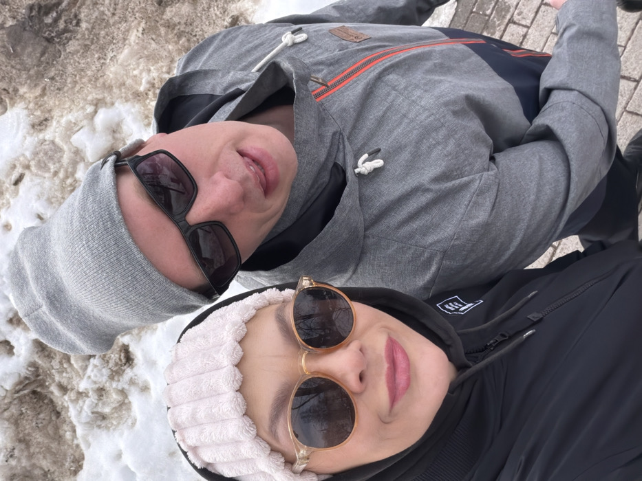 The image depicts two adults, an adult female and an adult male, posing for a close-up photograph outdoors in Chaikovsky, Russia. The female, positioned on the left, wears a black hooded jacket, a light pink ribbed headband, and round, light-framed sunglasses reflecting bare trees. Her lips are a light pink, and she exhibits a slight smile. The male, on the right, wears a gray beanie, black rectangular sunglasses, and a gray and dark blue color-block jacket with an orange zipper detail and a drawstring hood. His mouth is slightly open, with a portion of his tongue visible. The background consists of a snowy ground, partially melted and mixed with brown earth or dirt, suggesting a late winter or early spring environment. A paved surface is visible in the lower right foreground. Both individuals are looking towards the camera.