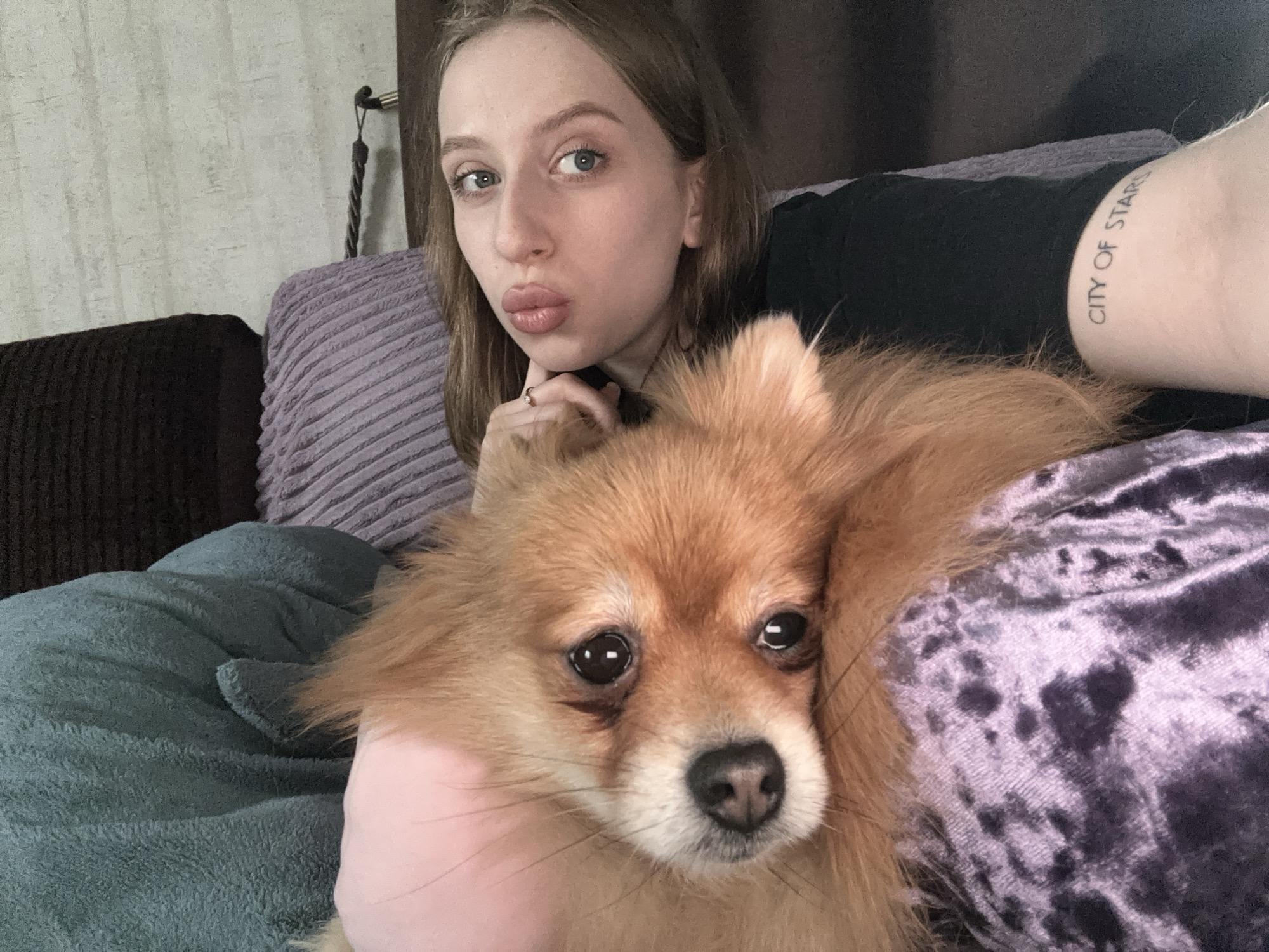 A full-shot selfie captures a young woman and a small, fluffy dog in a relaxed indoor setting, presumably in Chaikovsky, Russia. The woman, positioned towards the top of the frame, has light skin, light brown hair, and blue eyes, and is making a playful "kiss" face with pursed lips. She wears a black top and has a ring on her finger. A tattoo on her right arm is visible, reading "CITY OF STARS".
Cuddled closely with the woman is a reddish-brown dog, likely a Pomeranian, which occupies the lower and central part of the image. The dog looks directly at the camera with wide, dark, attentive eyes and a black nose, conveying a calm, slightly melancholic expression.
The subjects are surrounded by various textured fabrics: a light purple ribbed pillow behind the woman, a teal or blue plush blanket, and a dark purple crushed velvet material that the dog is resting upon. In the background, a light-colored, subtly textured wallpaper covers the wall, with a black cord or strap hanging visible on the left. The scene evokes a cozy, domestic atmosphere during what appears to be daytime.