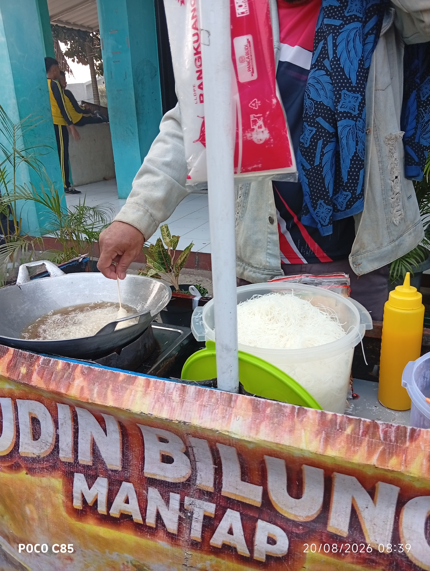 The image depicts an outdoor food stall in South Tangerang, Indonesia. A person is frying food in a large metal wok filled with oil. A large white plastic container filled with white noodles is visible next to the wok. A yellow squeeze bottle, likely for sauce or condiments, is also present. In the background, two young males in yellow and black athletic wear are visible. The food stall has a banner with text that reads "UDIN BILUNG MANTA" and "POCO C85" and a date and time stamp "20/08/2026 08:39". The overall scene suggests a busy food preparation environment, possibly at a local market or street food vendor.