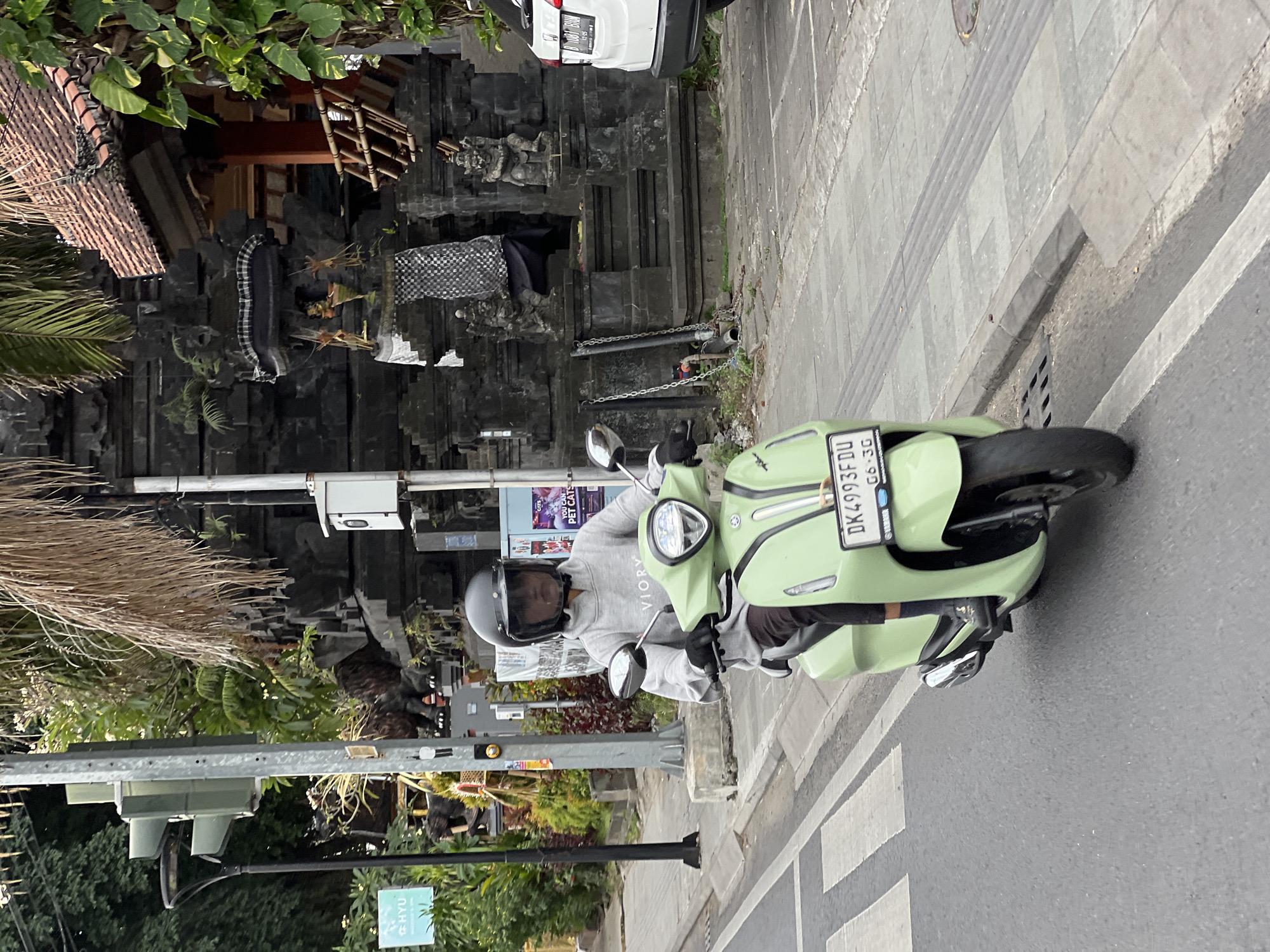 The image shows a person riding a light green scooter on a street in Denpasar, Indonesia. The rider is wearing a dark helmet and a grey hooded sweatshirt. The scooter has a license plate that reads "DK4993FDU". In the background, there is a traditional Balinese building with intricate stone carvings and a thatched roof. A traffic light is visible on the left side of the image. A white car is parked on the right side of the street. The street has a pedestrian crossing and a sidewalk. The scene appears to be a typical street view in Denpasar, a city known for its vibrant culture and religious heritage.