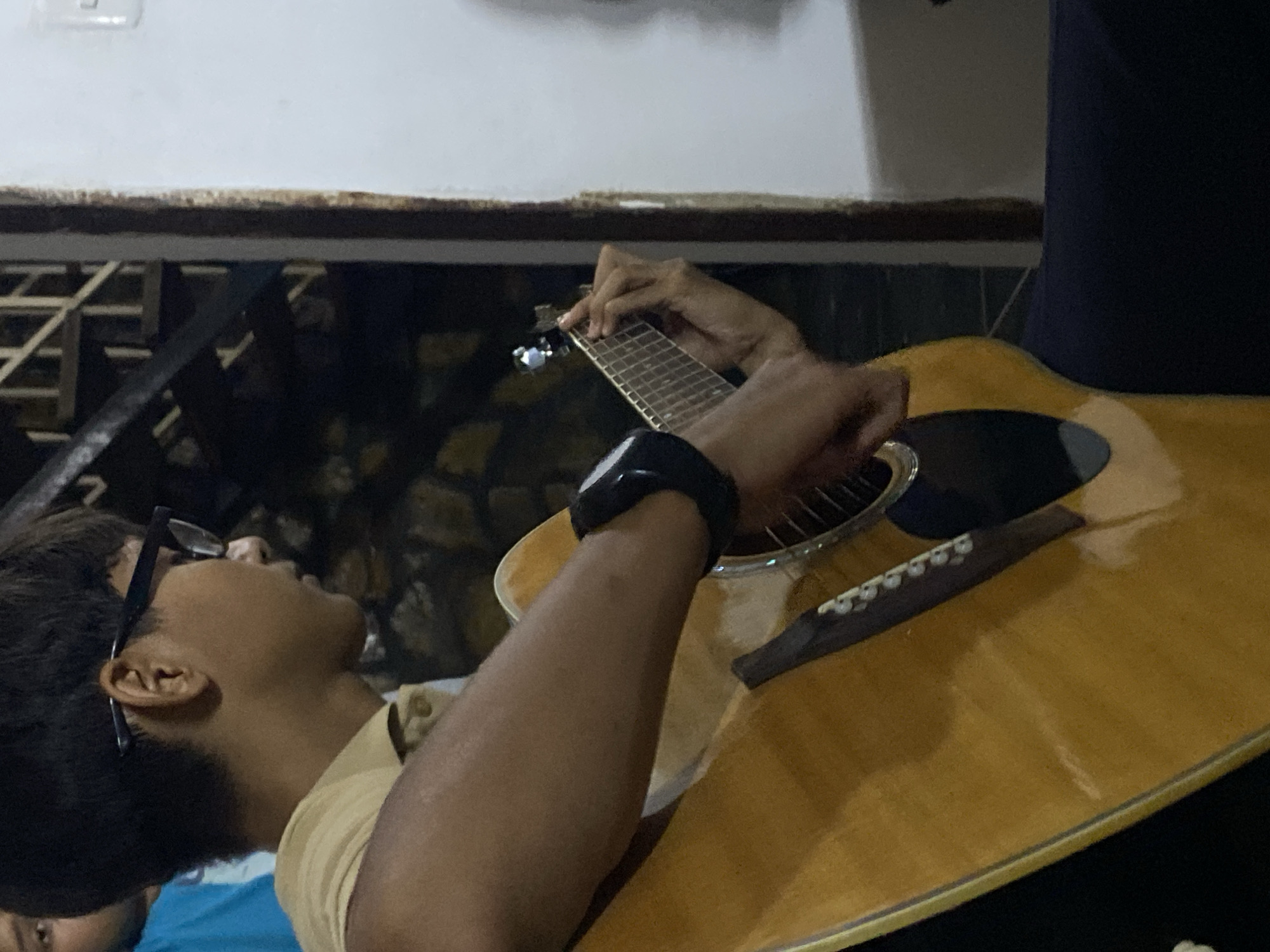 A person is playing an acoustic guitar. The individual wears glasses and a black watch. The setting appears to be indoors, possibly a home or building in Tinaquillo, Venezuela. The background includes a wall and what seems to be a window or railing.