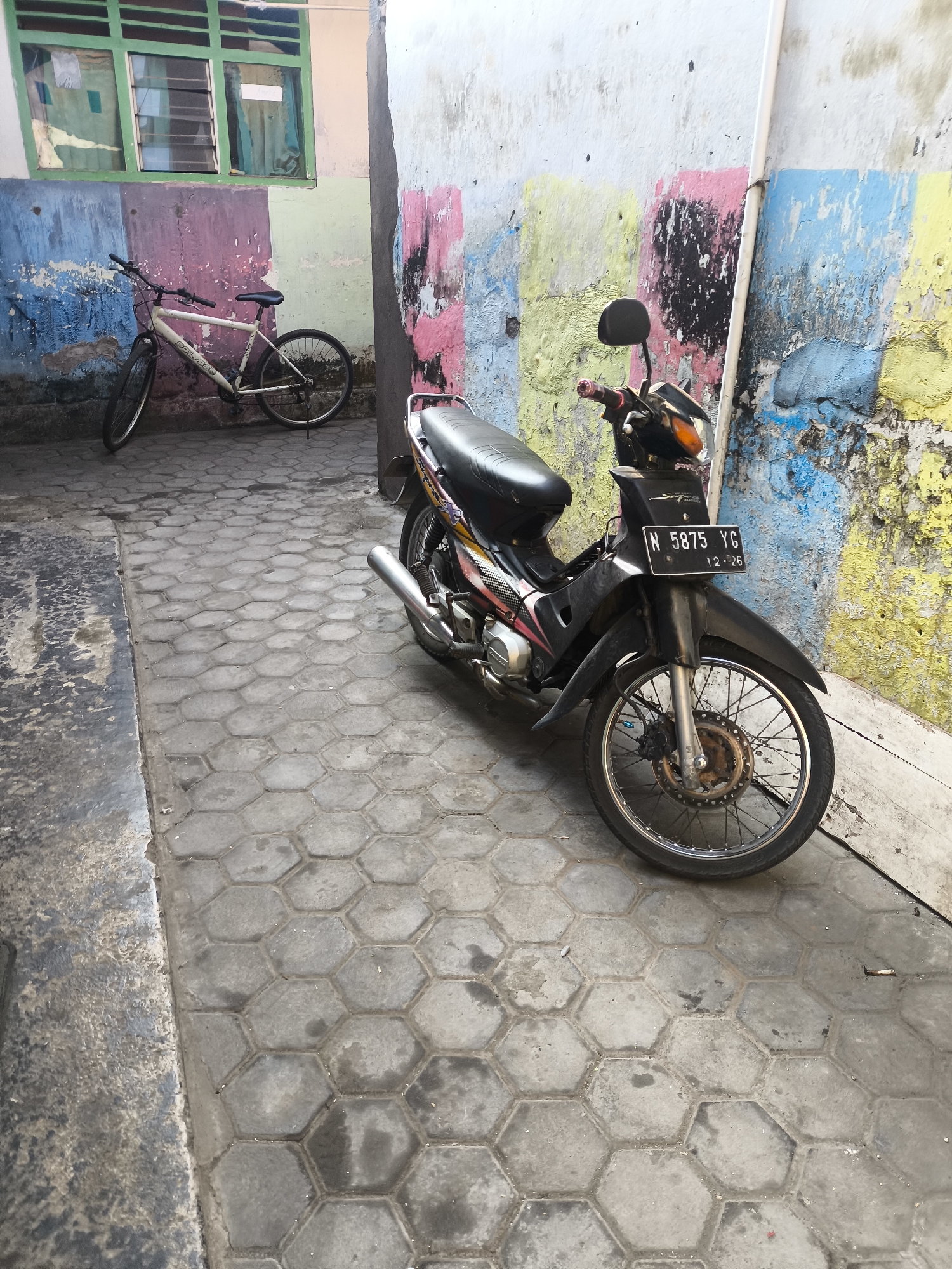 An outdoor medium shot captures a narrow, tiled alleyway. A black and silver motorcycle is parked on the right side of the alley, facing forward. The motorcycle has a license plate that reads "N 5875 YG" with a date of "12-26". To the left of the motorcycle, a white bicycle is parked against a wall adorned with colorful, vertical stripes of blue, purple, pink, and yellow. The wall also features a green-framed window with a faded curtain visible inside. The ground is paved with grey hexagonal tiles. The scene appears to be in a city environment, likely in a residential or commercial area, given the presence of vehicles and the colorful painted wall which suggests an attempt at beautification or artistic expression. The lighting indicates it is daytime, casting some shadows but generally illuminating the scene. The image is taken from a slightly low angle, looking into the alleyway.